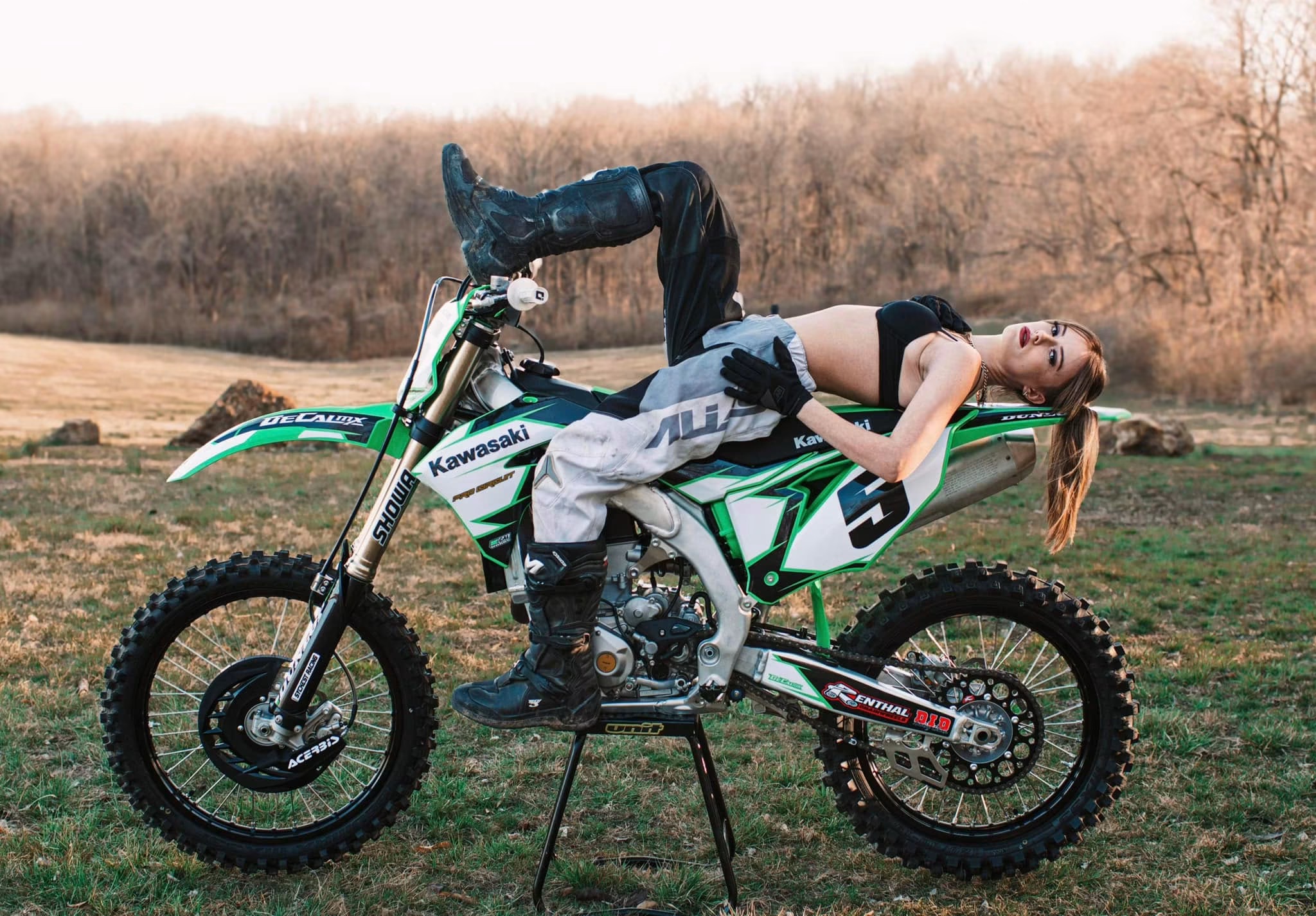 Person lying on a dirt bike outdoors, with legs and head hanging over the sides, in a grassy field.