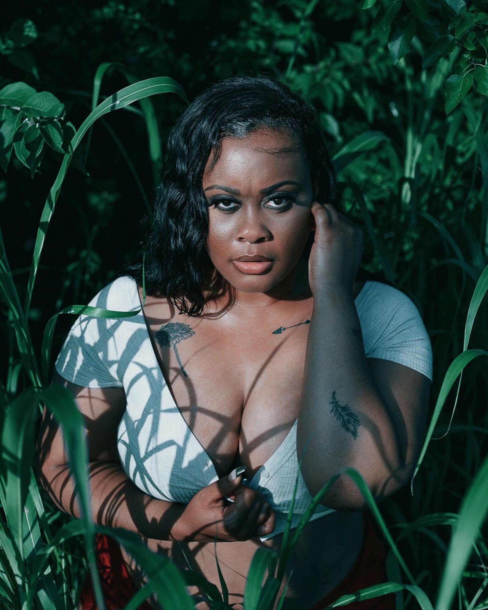 Woman with dark hair and tattoos among tall green plants, looking at the camera with a serious expression.