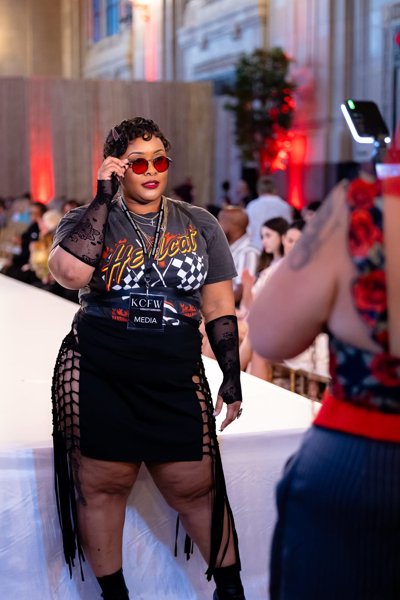 Person with curly hair wearing sunglasses, a black dress with fishnet details, and gloves, posing at an event.