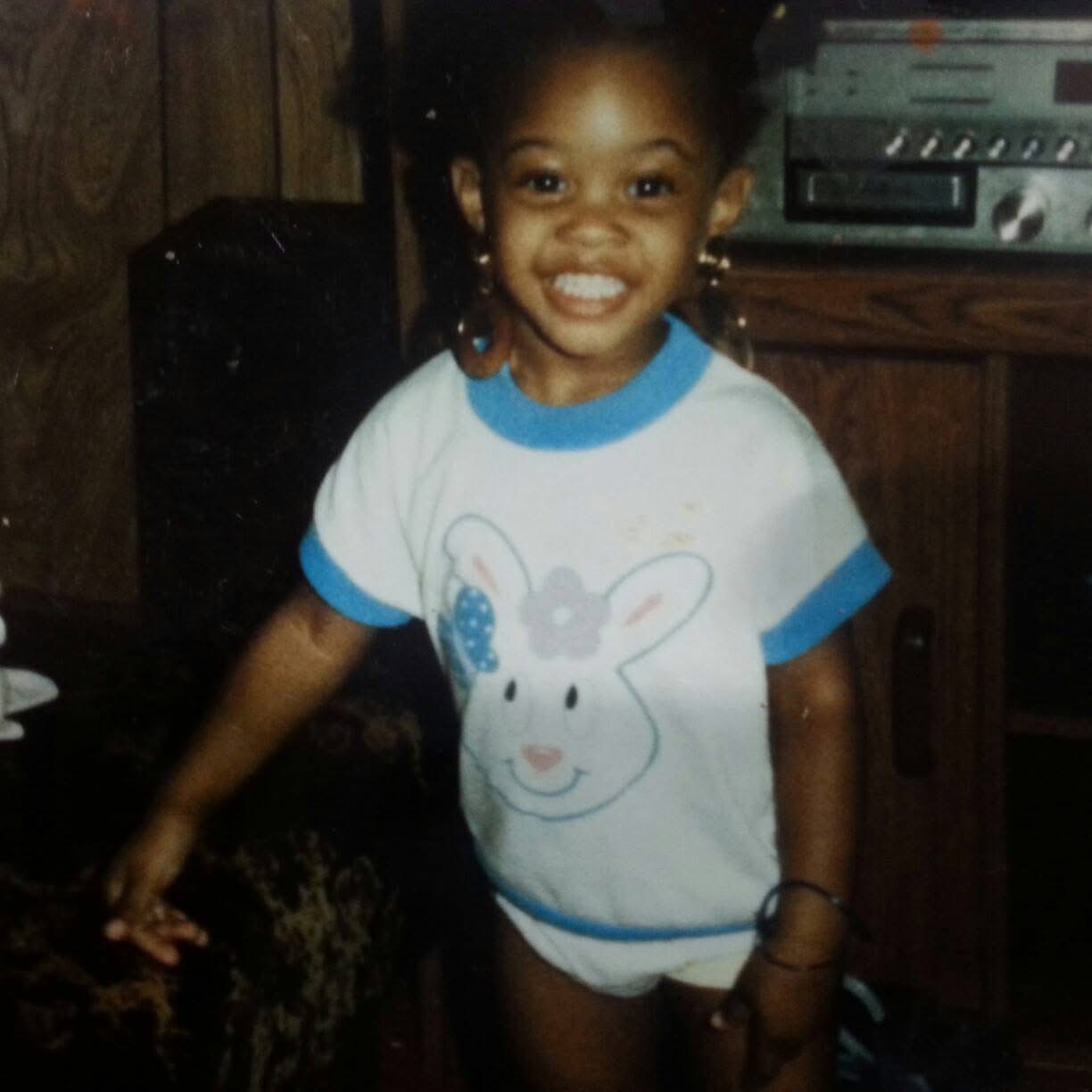 Young girl smiling, wearing a white T-shirt with a bunny face, in a room with wooden furniture and electronic equipment.