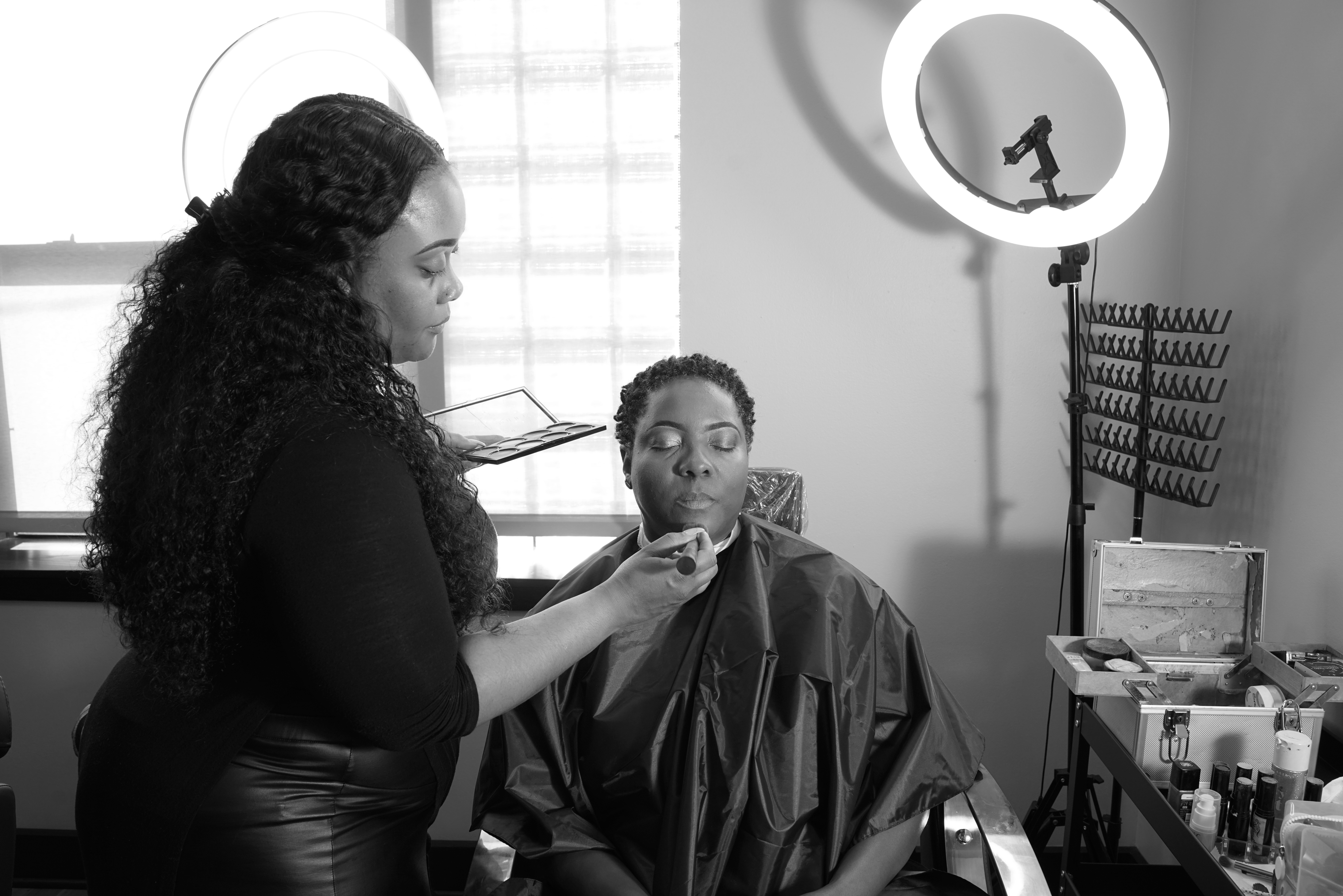 Makeup artist applies makeup to a person seated in a salon chair, with ring light and makeup tools visible.