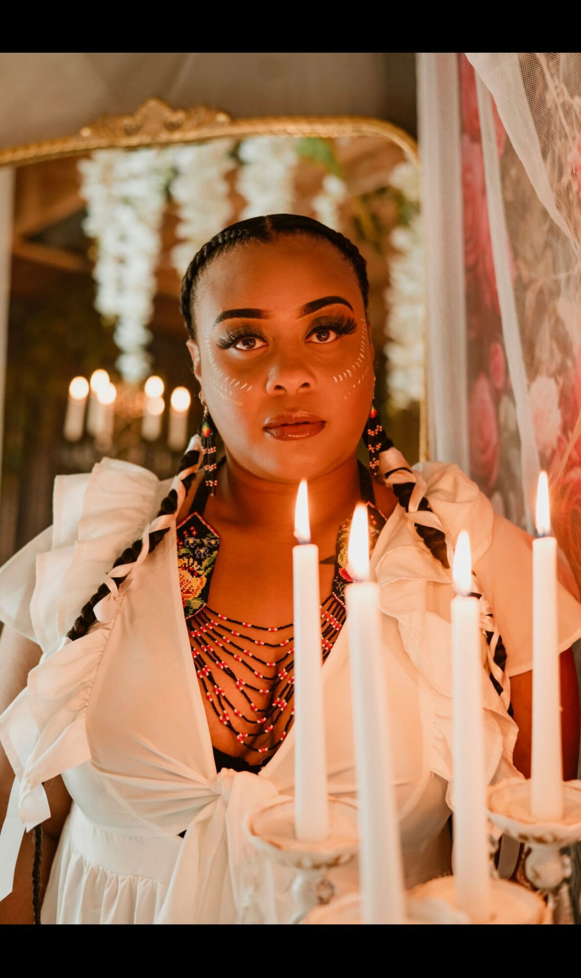 Woman with braided hair and makeup, surrounded by lit candles, in a decorated indoor setting.