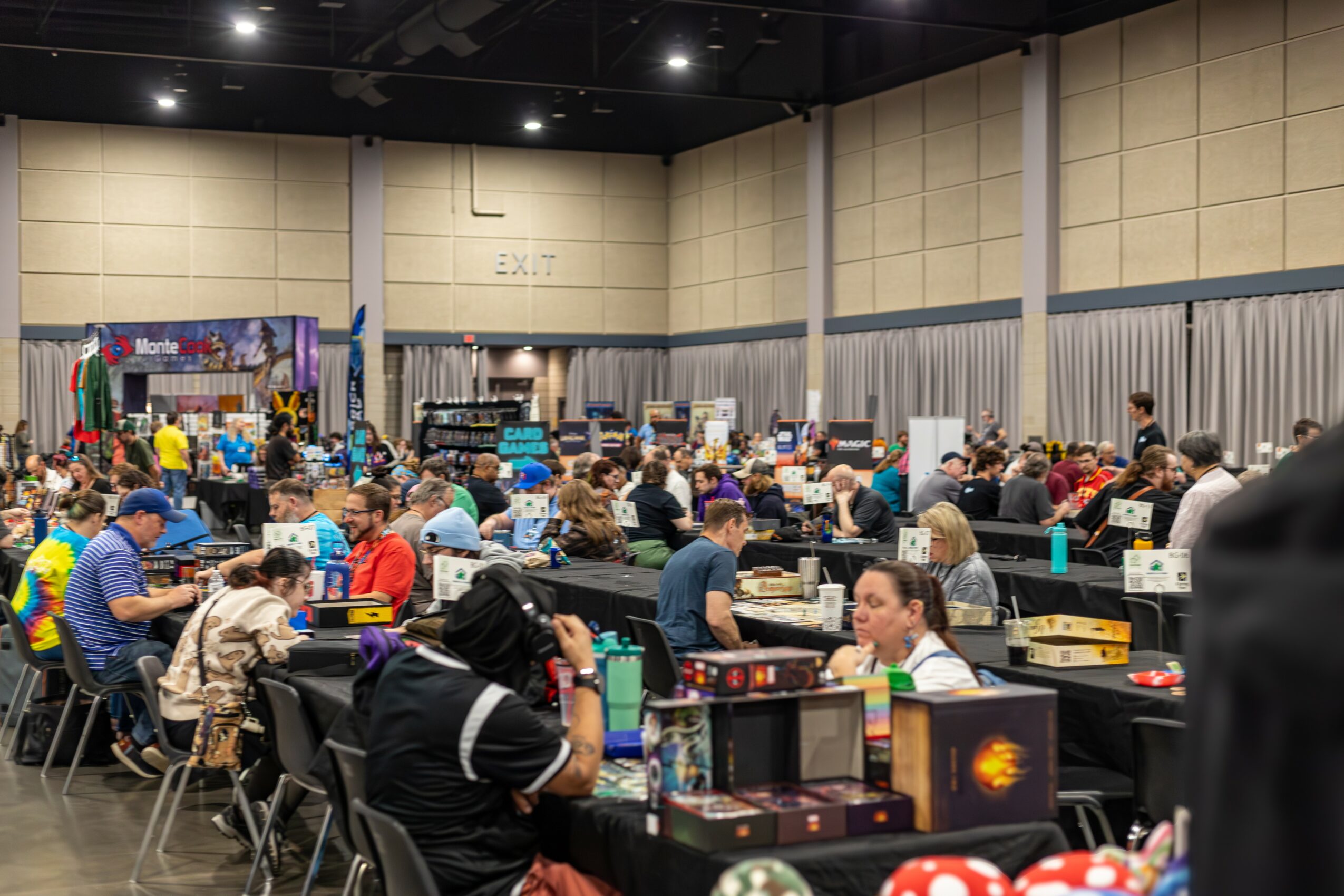 Large indoor event with many people seated at tables, engaging in activities, with booths and displays in the background.