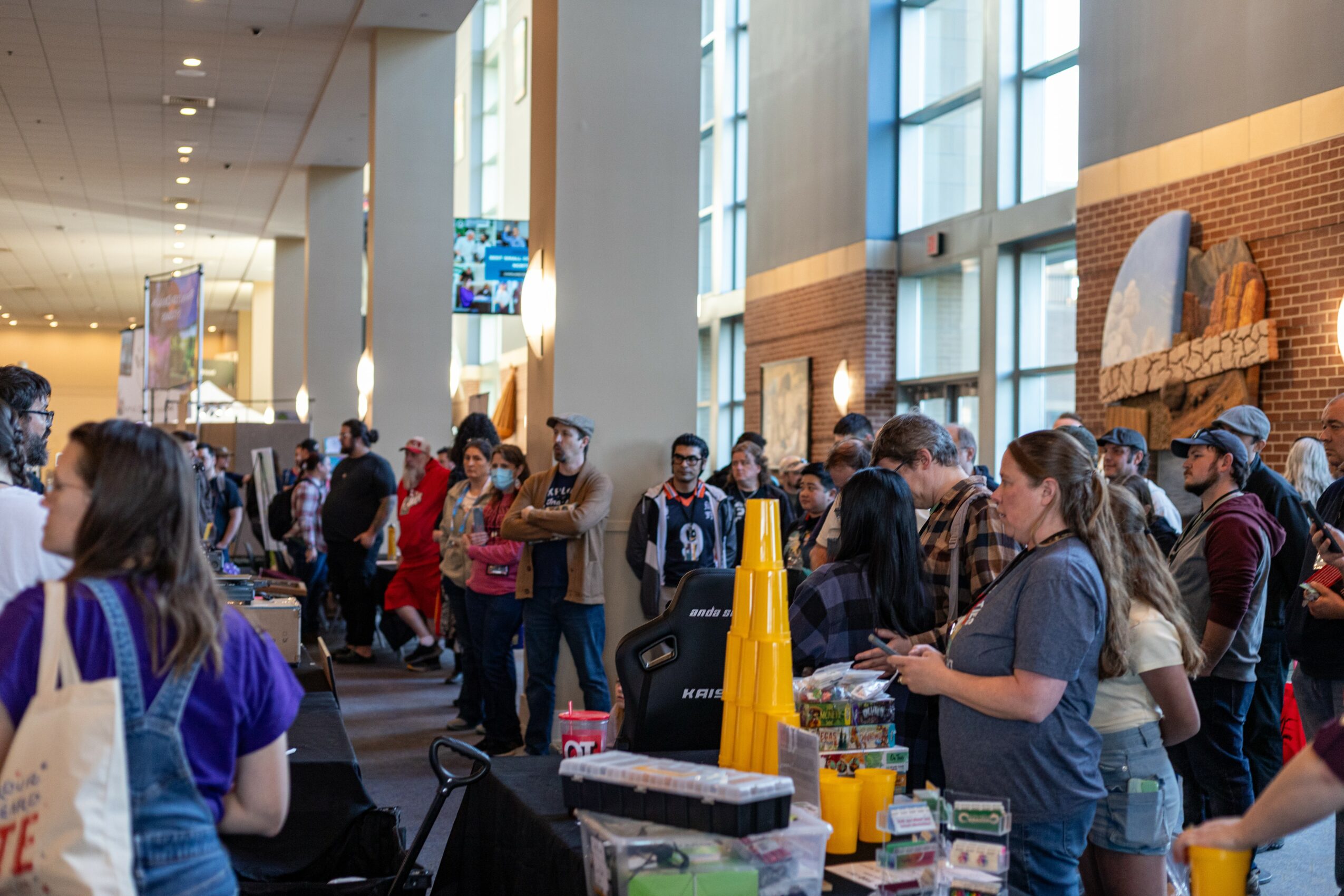 People gather in a spacious indoor area with large windows, some standing in line or talking, and a woman serving drinks.