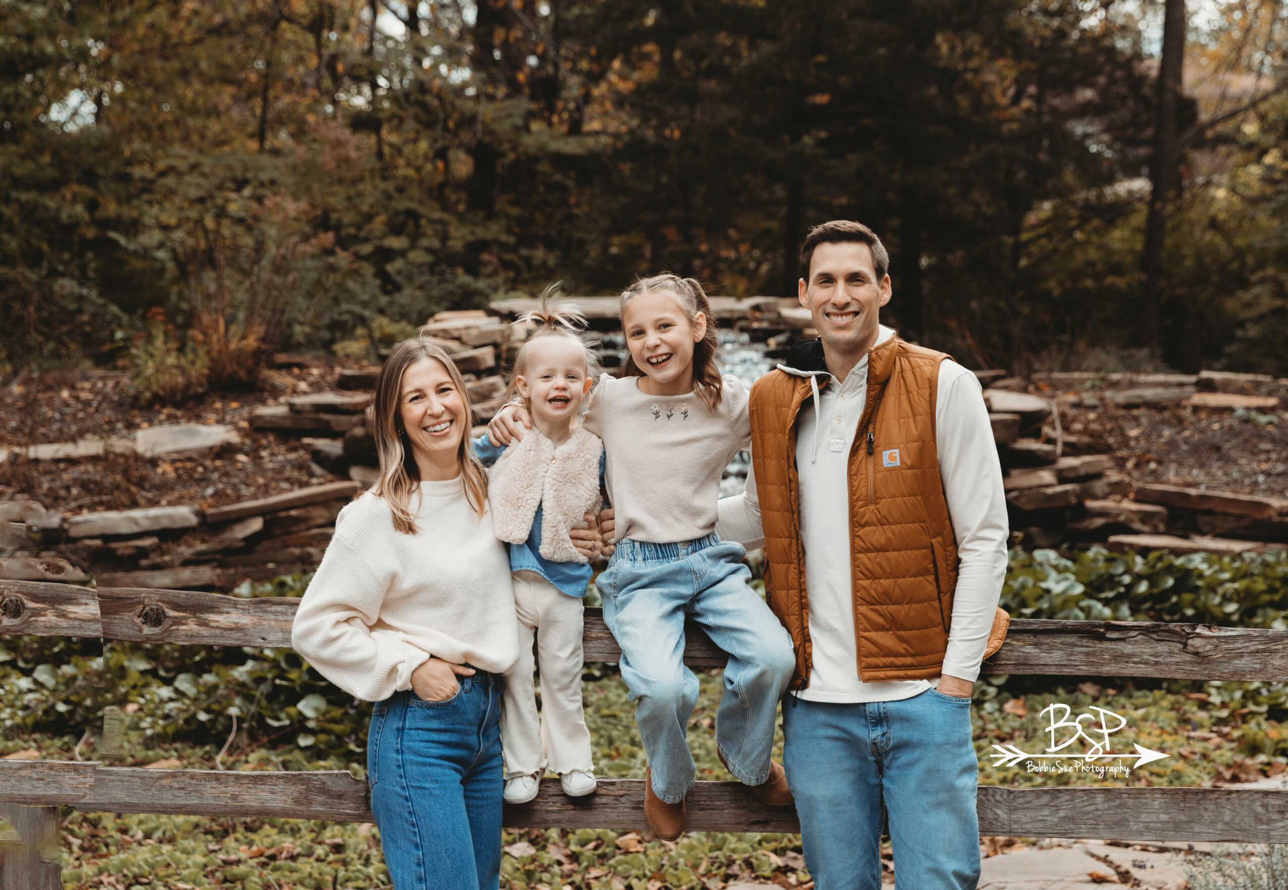 Four people, two children and two adults, standing outdoors in front of a wooden fence and trees, smiling.