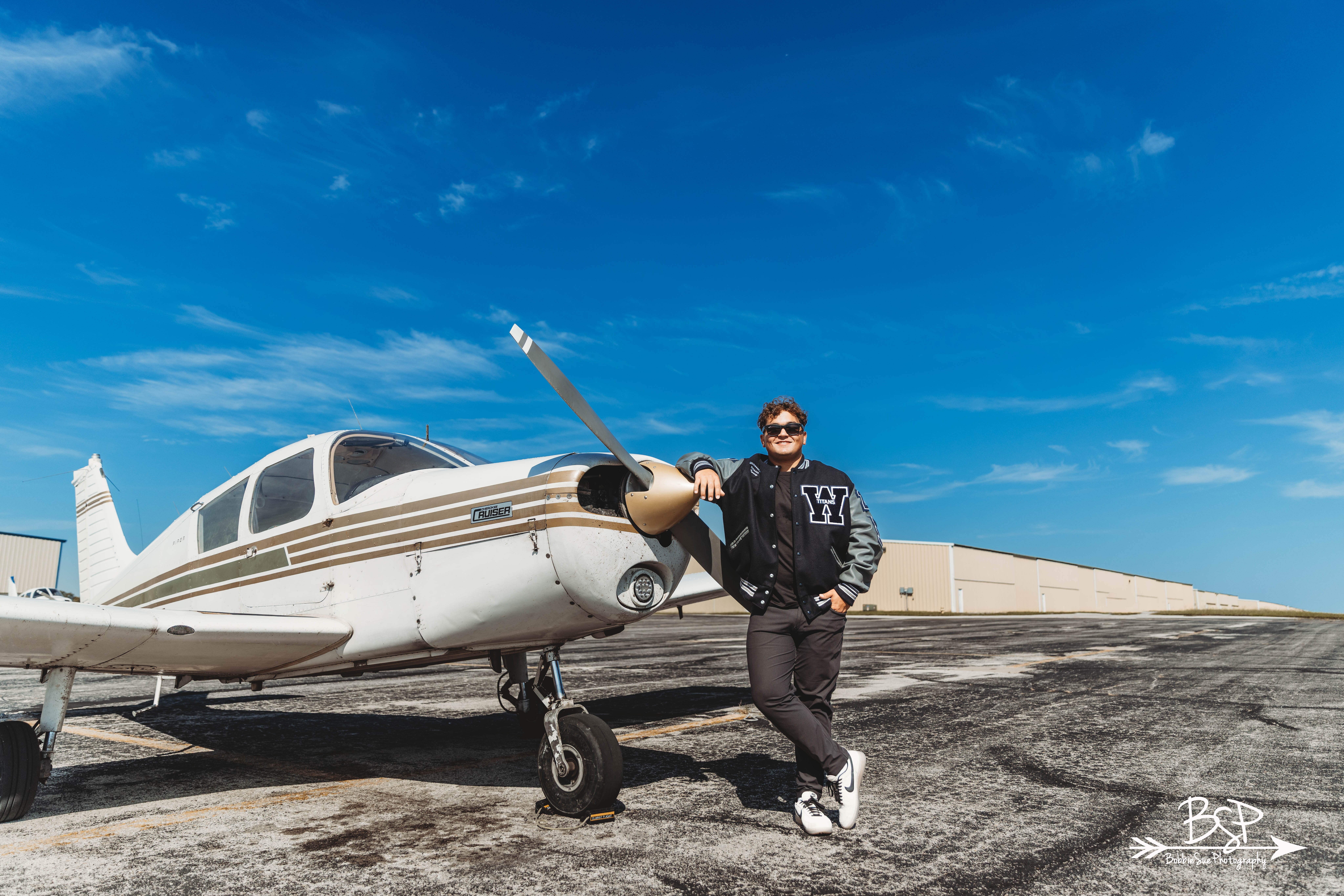 Person standing next to small airplane on tarmac under blue sky with clouds, wearing sunglasses and casual clothing.