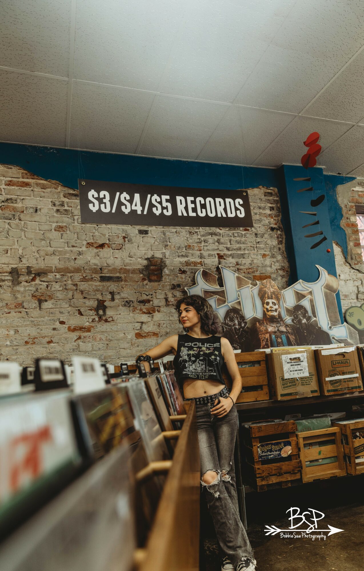 Young woman with dark hair, wearing a black crop top and ripped jeans, stands in record store with brick wall background.