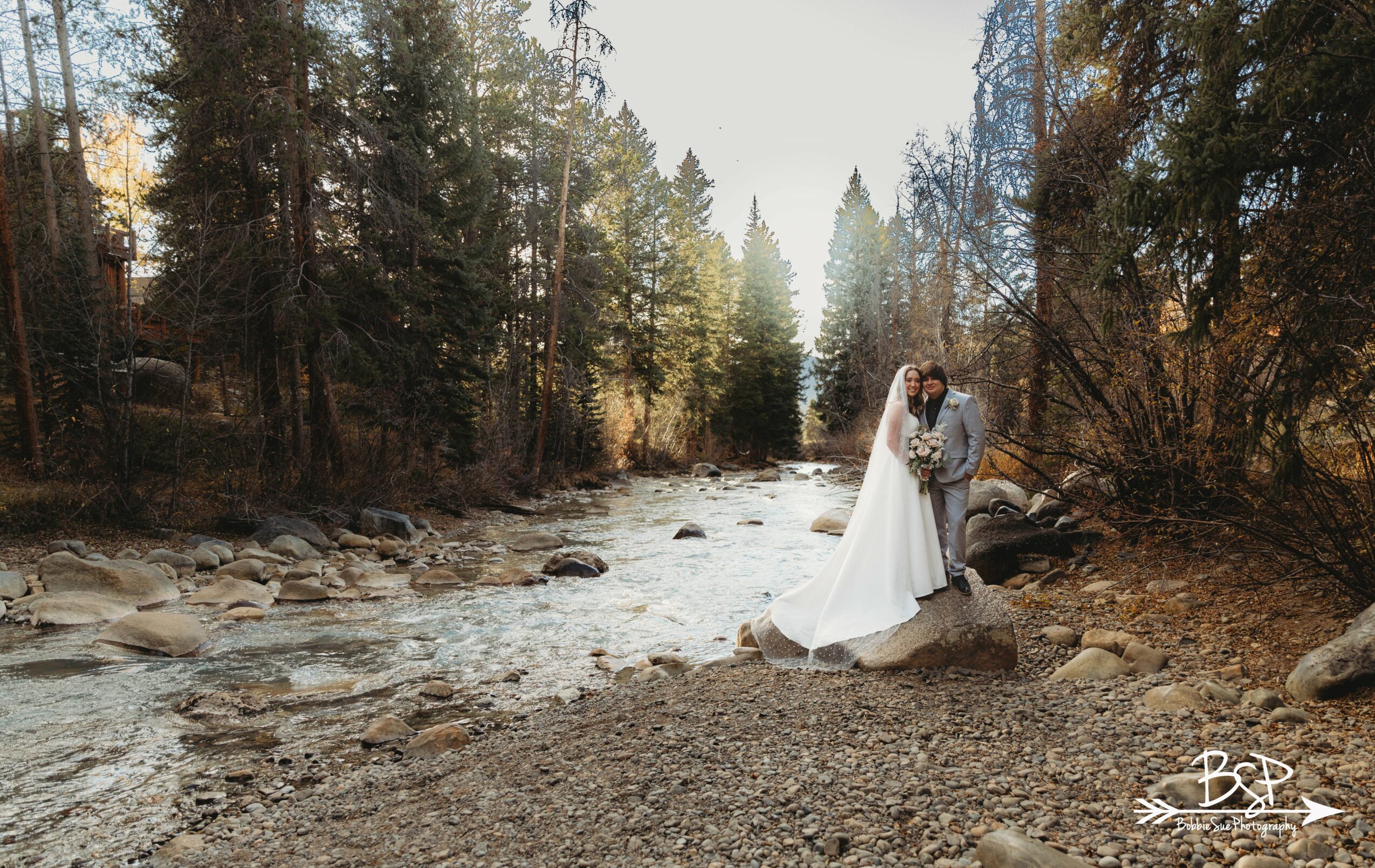 A couple in wedding attire standing by a river in a forested area, with trees and rocks around them.