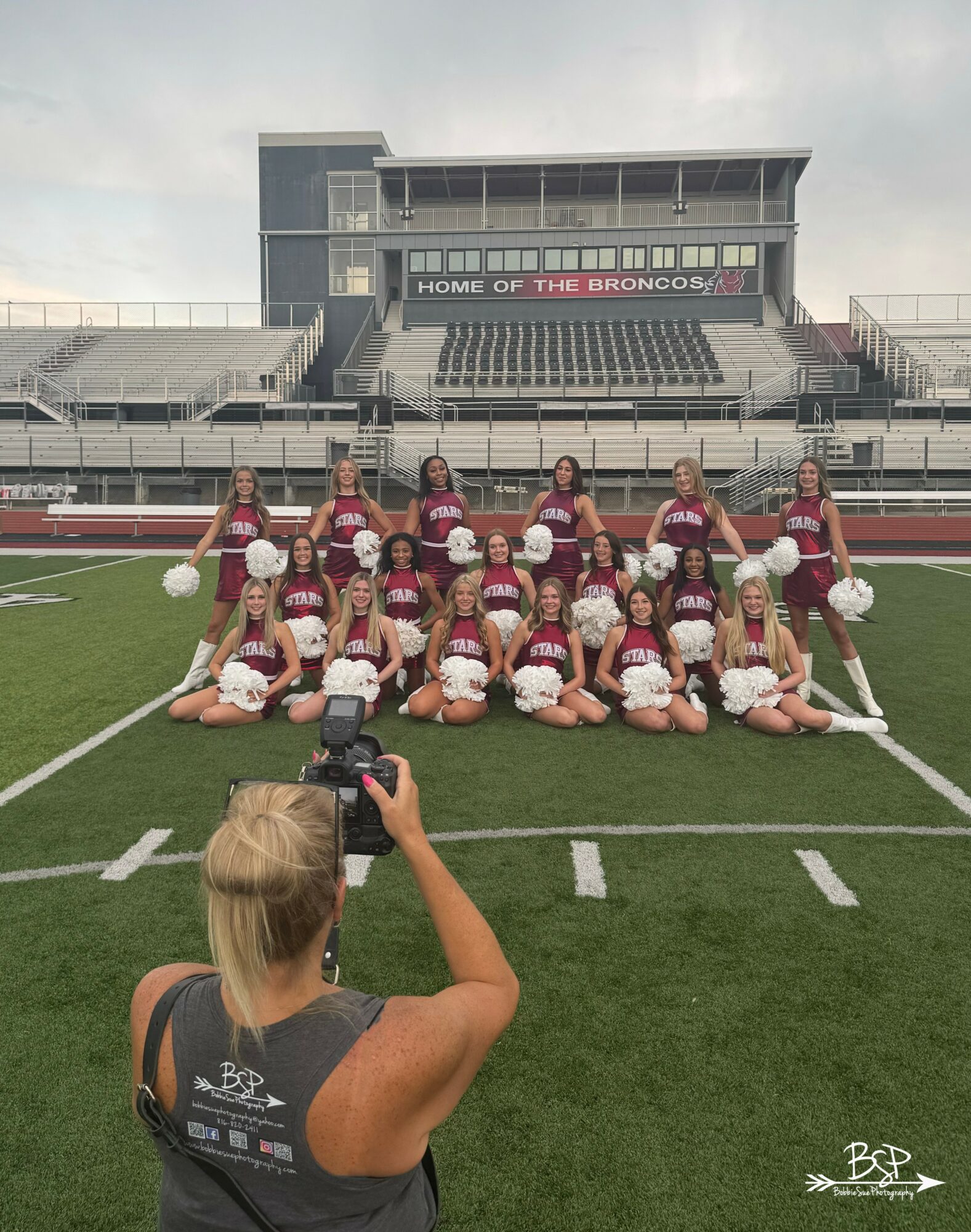 Group of cheerleaders in maroon uniforms holding white pom-poms on a football field, with a stadium and scoreboard in background.