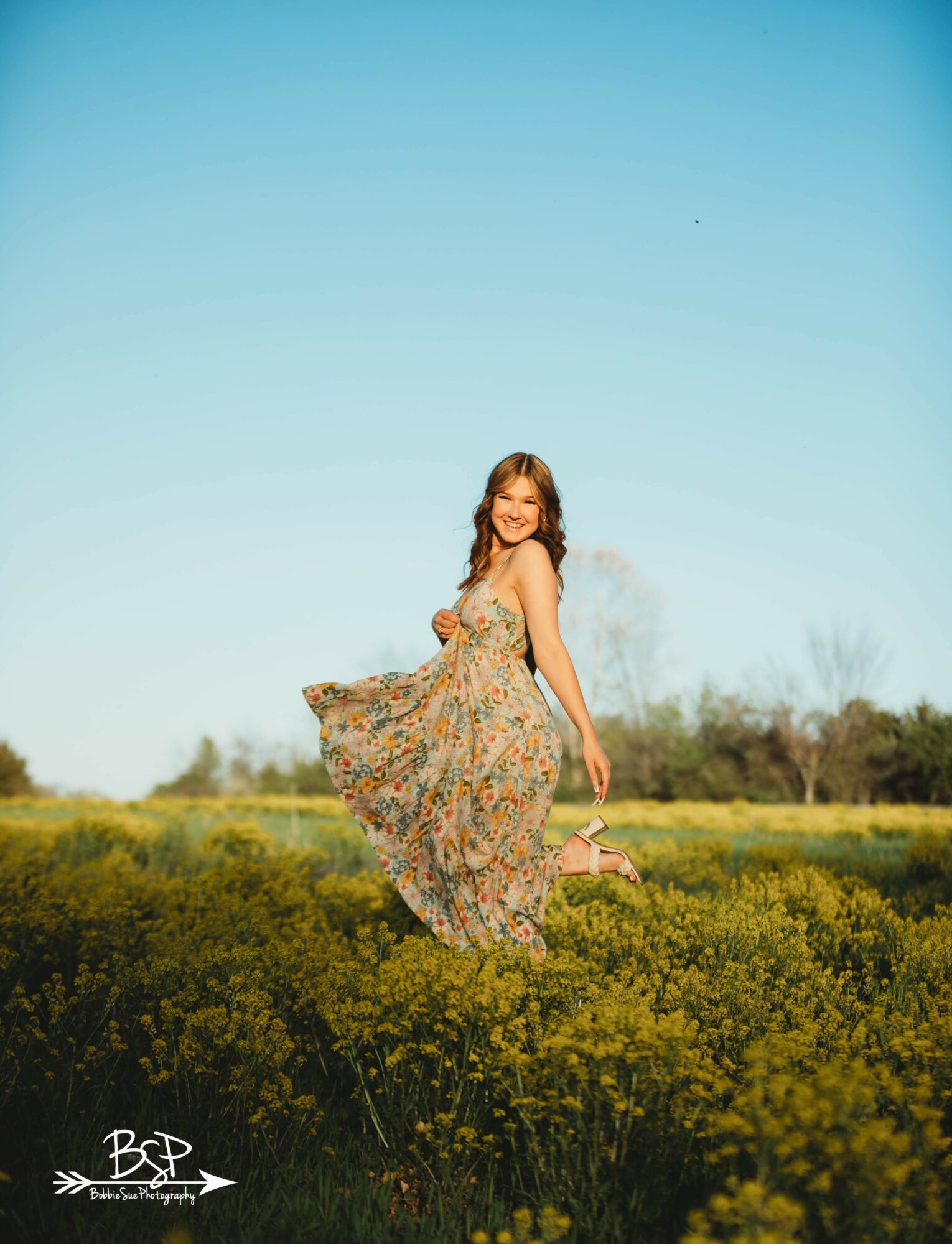 Young woman in a floral dress standing in a field of yellow flowers under a clear blue sky, smiling and looking back.