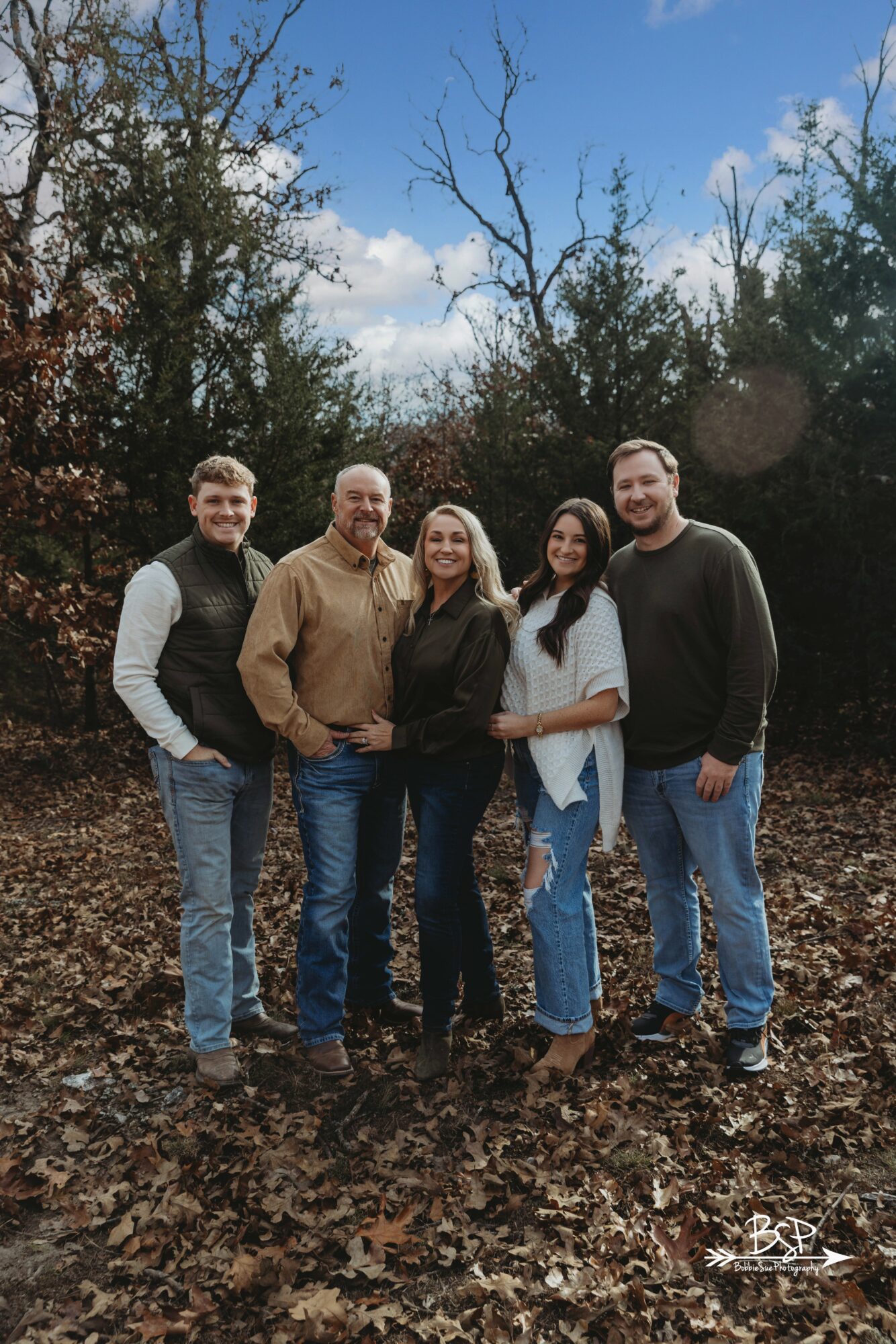 Group of five people standing outdoors on fallen leaves, trees, and blue sky in background.