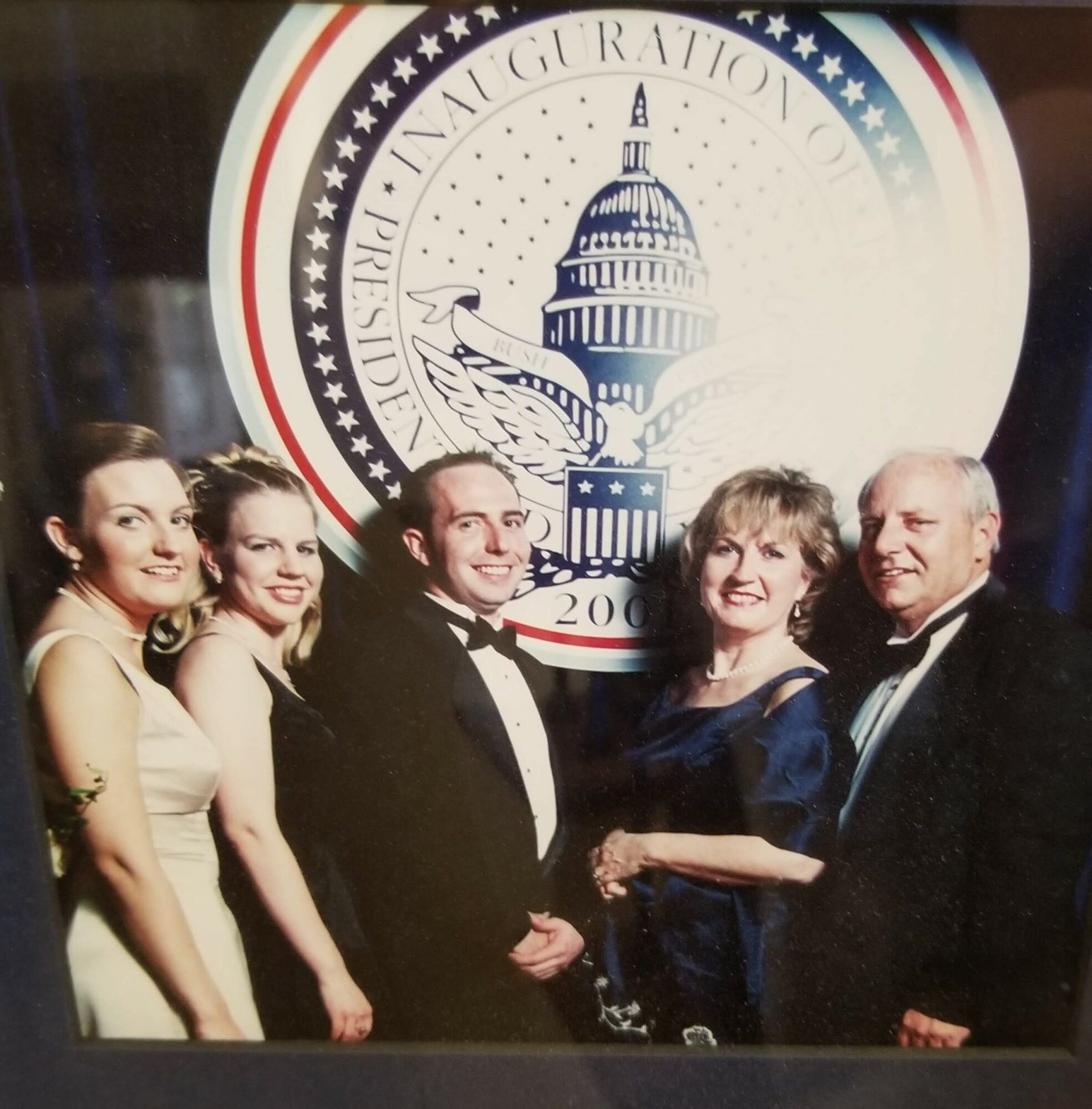 Group of five people in formal attire standing in front of a large circular emblem with a dome and eagle, smiling.