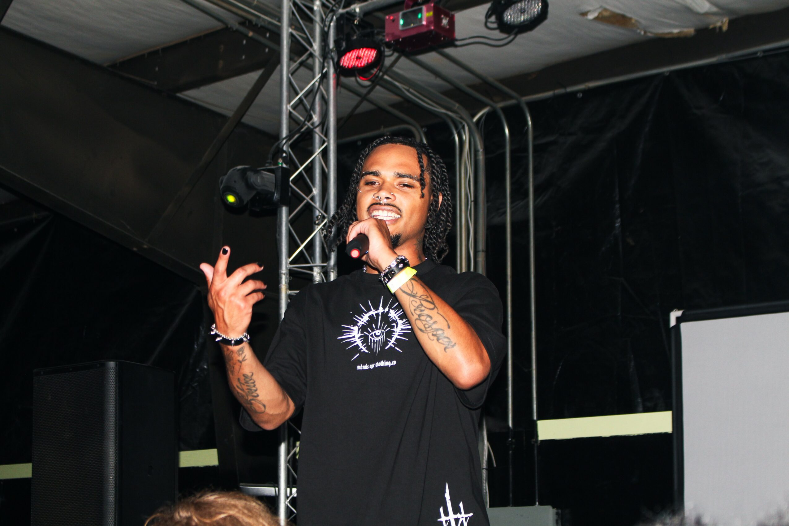 Person with long hair smiling and holding a microphone on stage, wearing a black T-shirt with a design, in front of stage equipment.