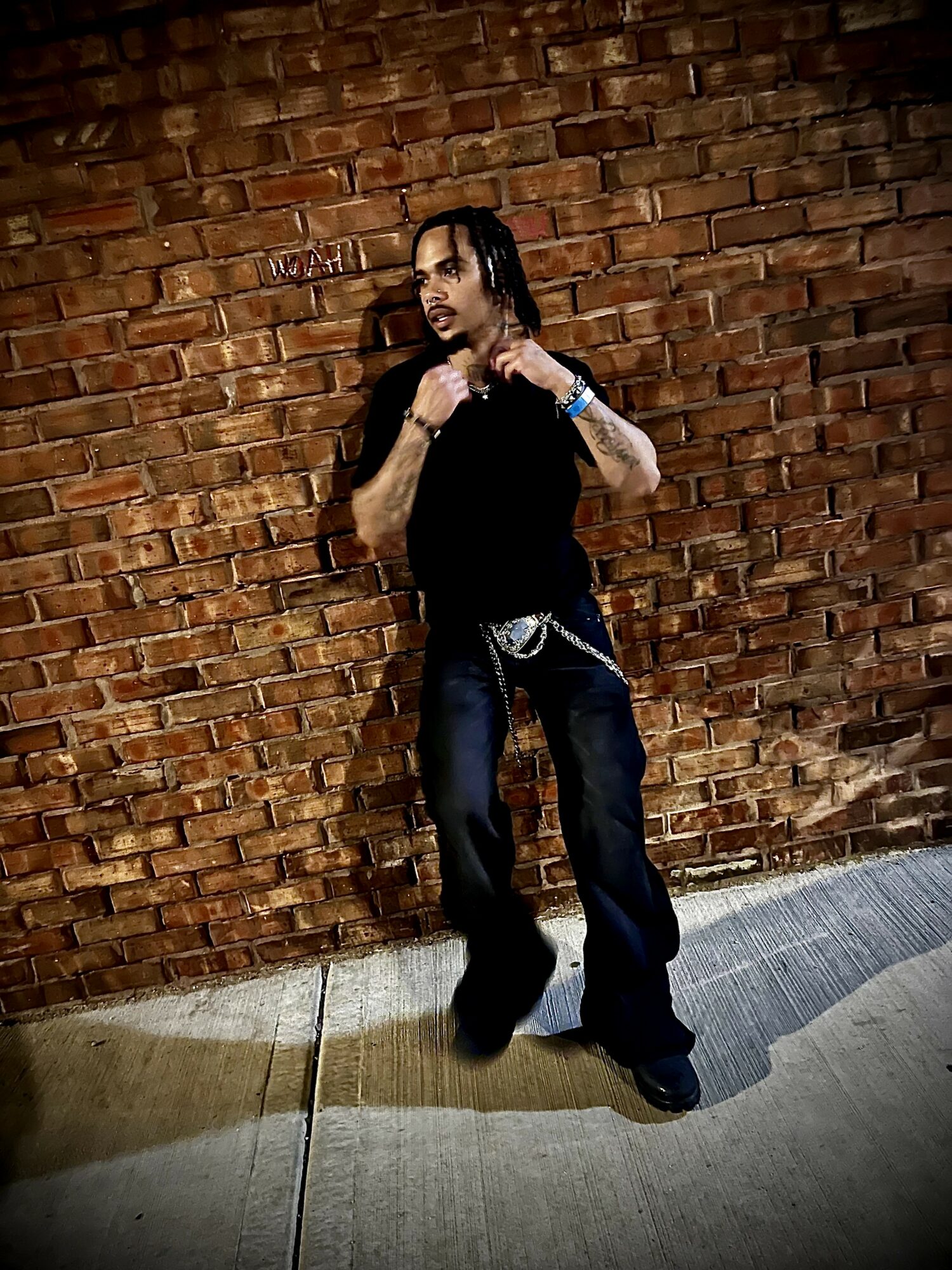 Person with long hair and dark clothing standing against a brick wall, looking at the camera.