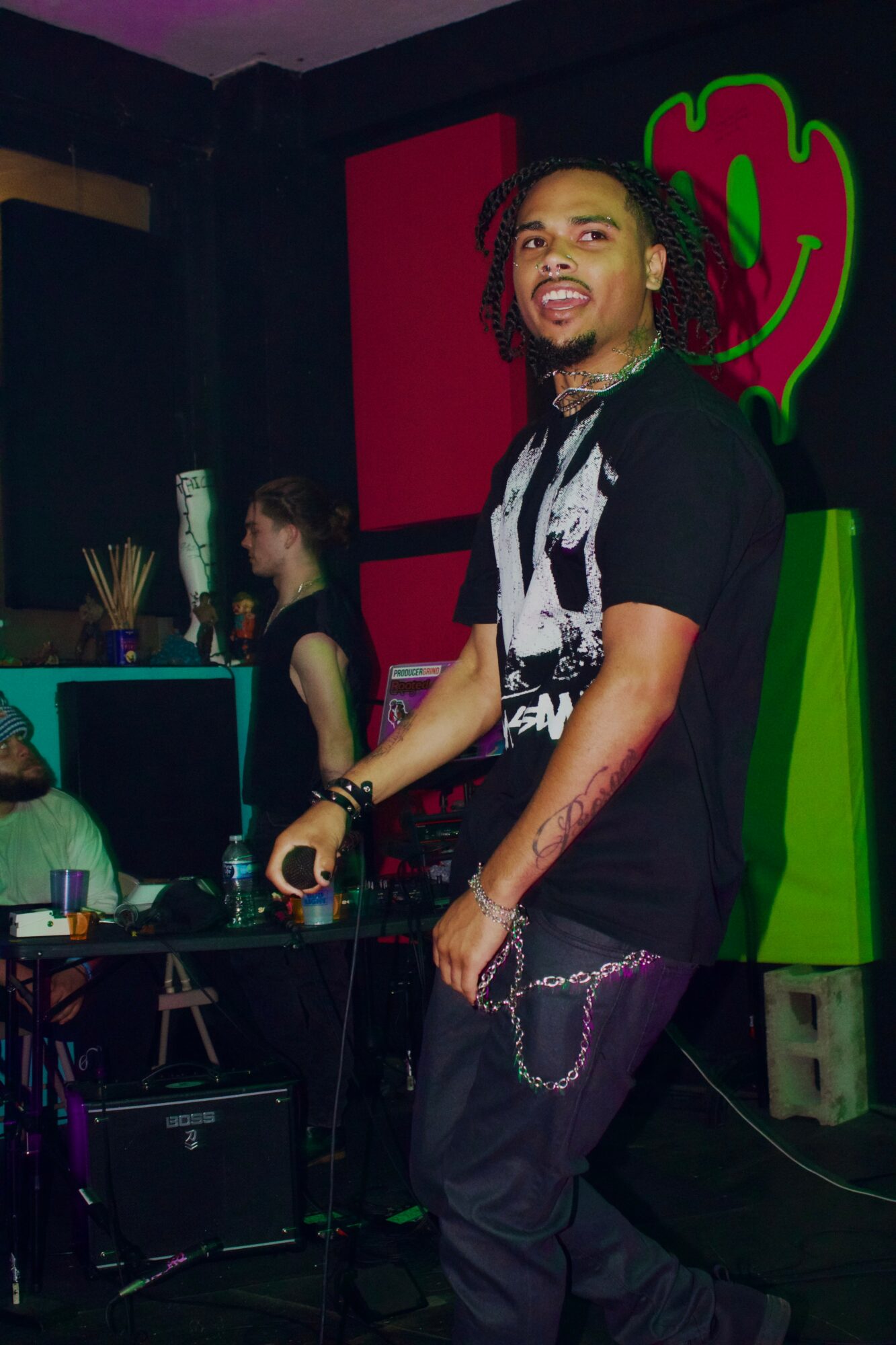 Person with dreadlocks and tattoos standing in a club with colorful background, holding a microphone, smiling.