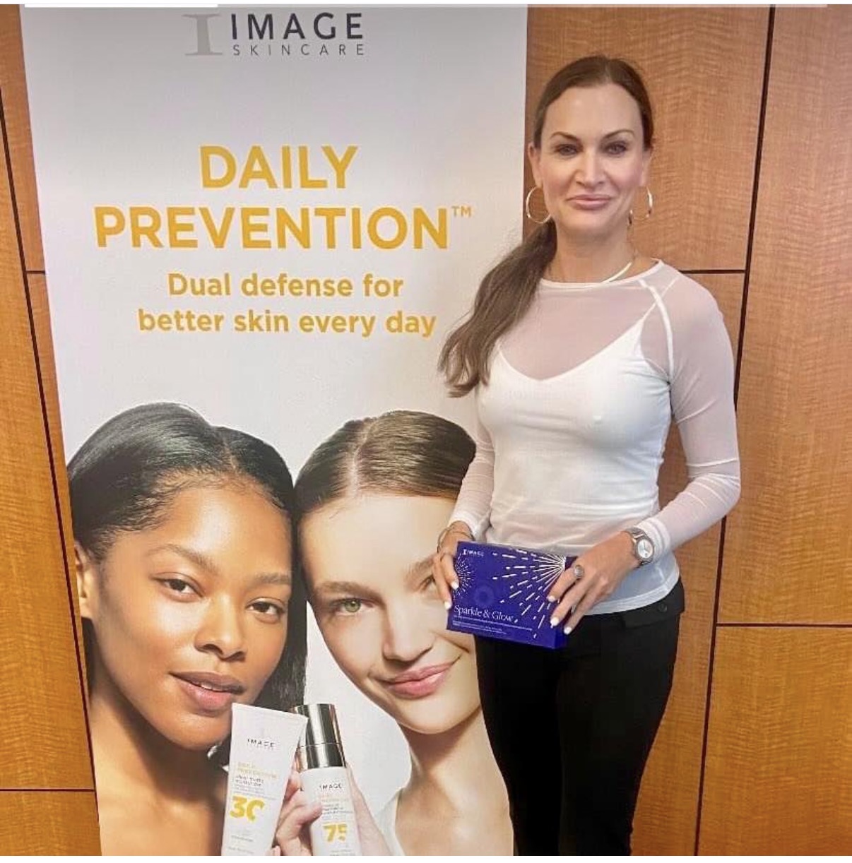 Woman standing next to a skincare poster, holding a product, smiling, in an indoor setting.