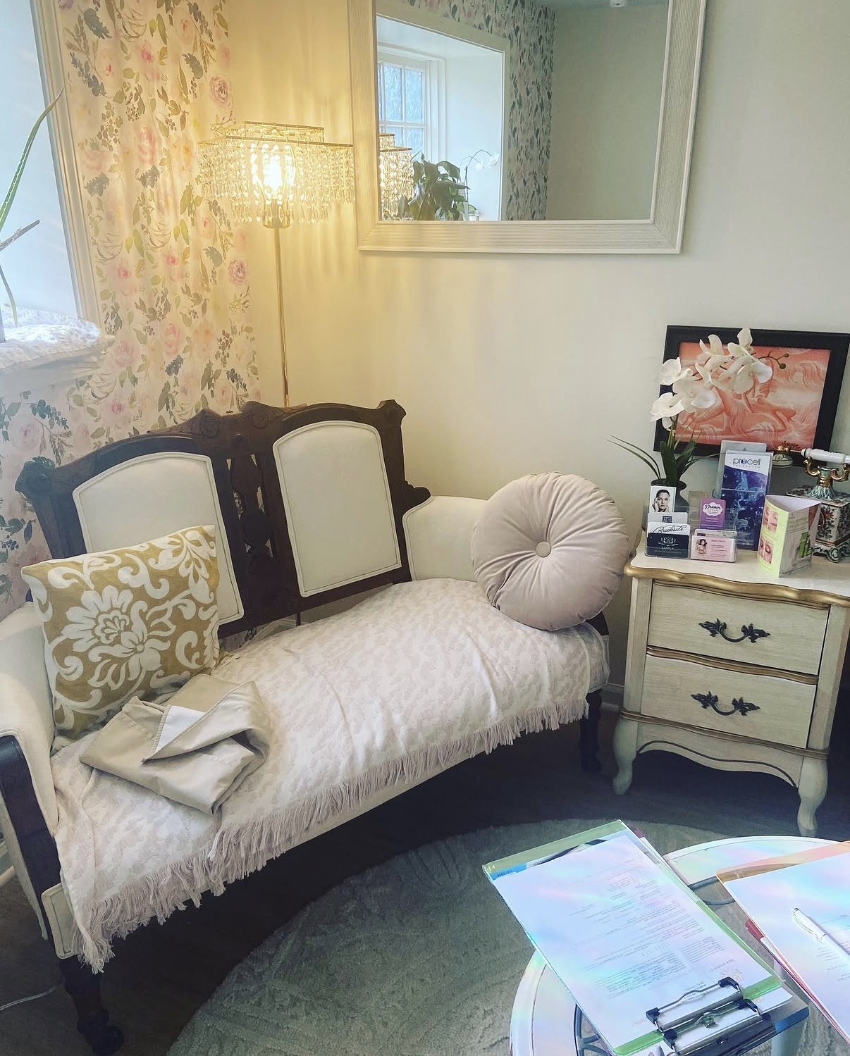 A vintage-style sofa with pillows and a blanket, a side table with items, a mirror, and a window with floral wallpaper.