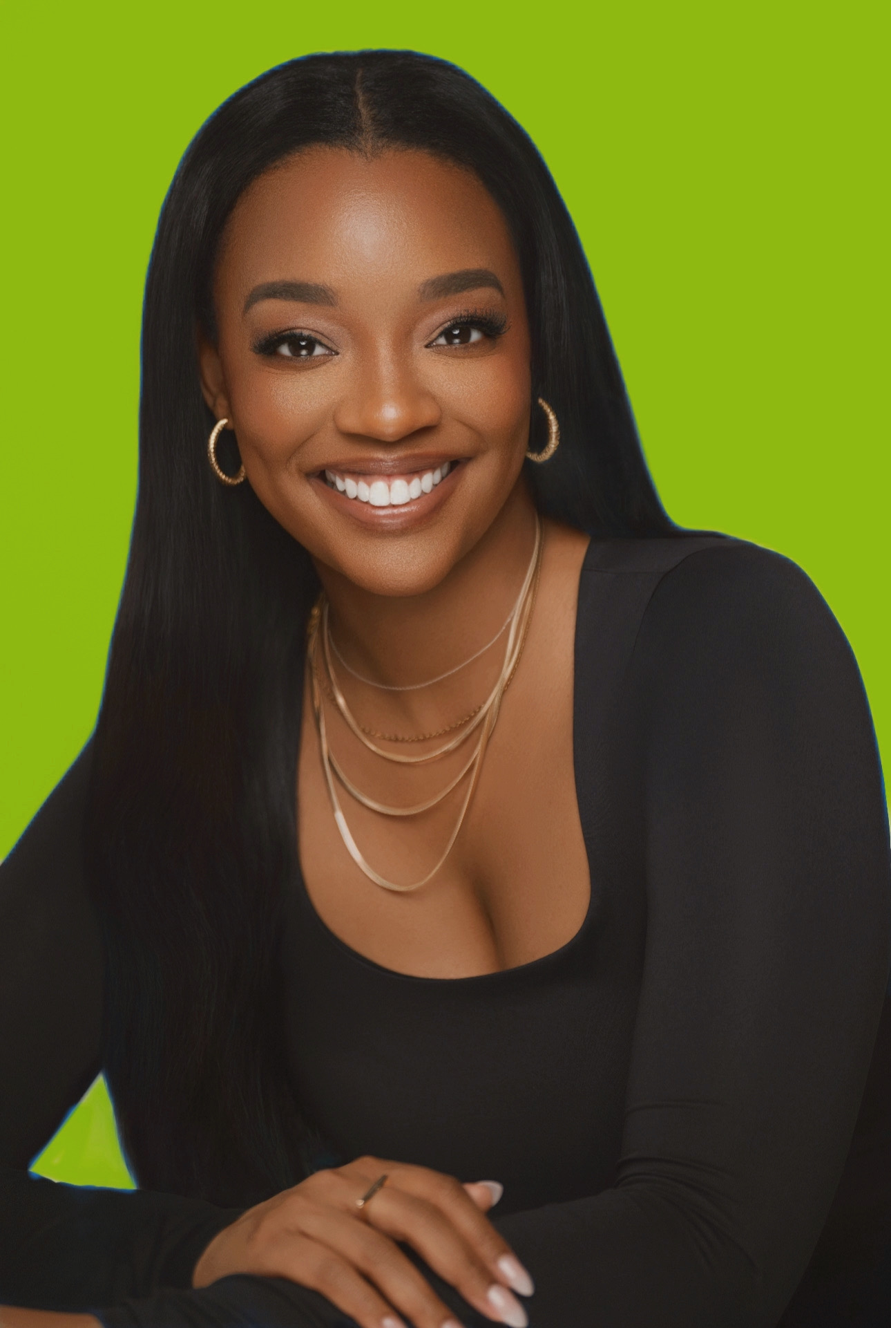 Smiling woman with long black hair, wearing gold jewelry and a black top, against a green background.