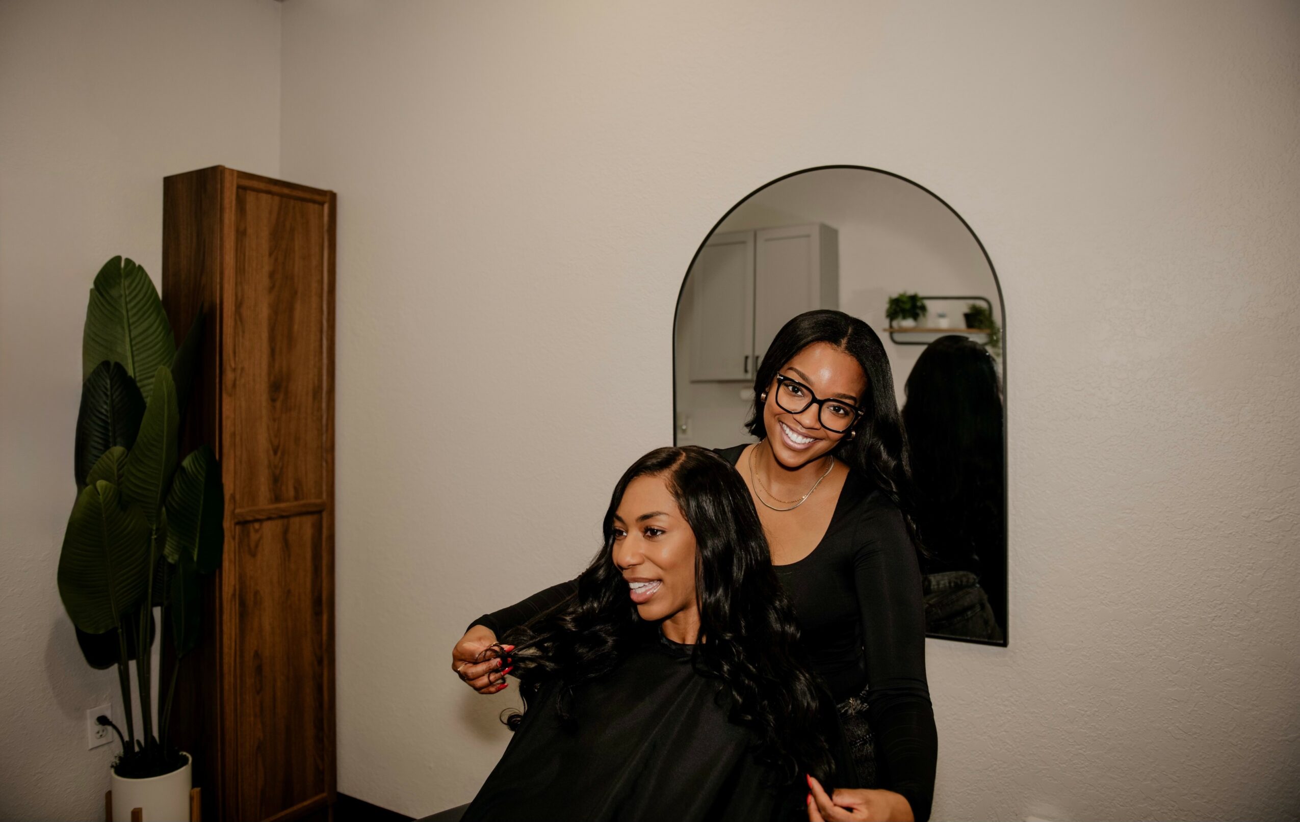 Two women smiling, one sitting and the other standing, in a room with a mirror and a wooden cabinet.