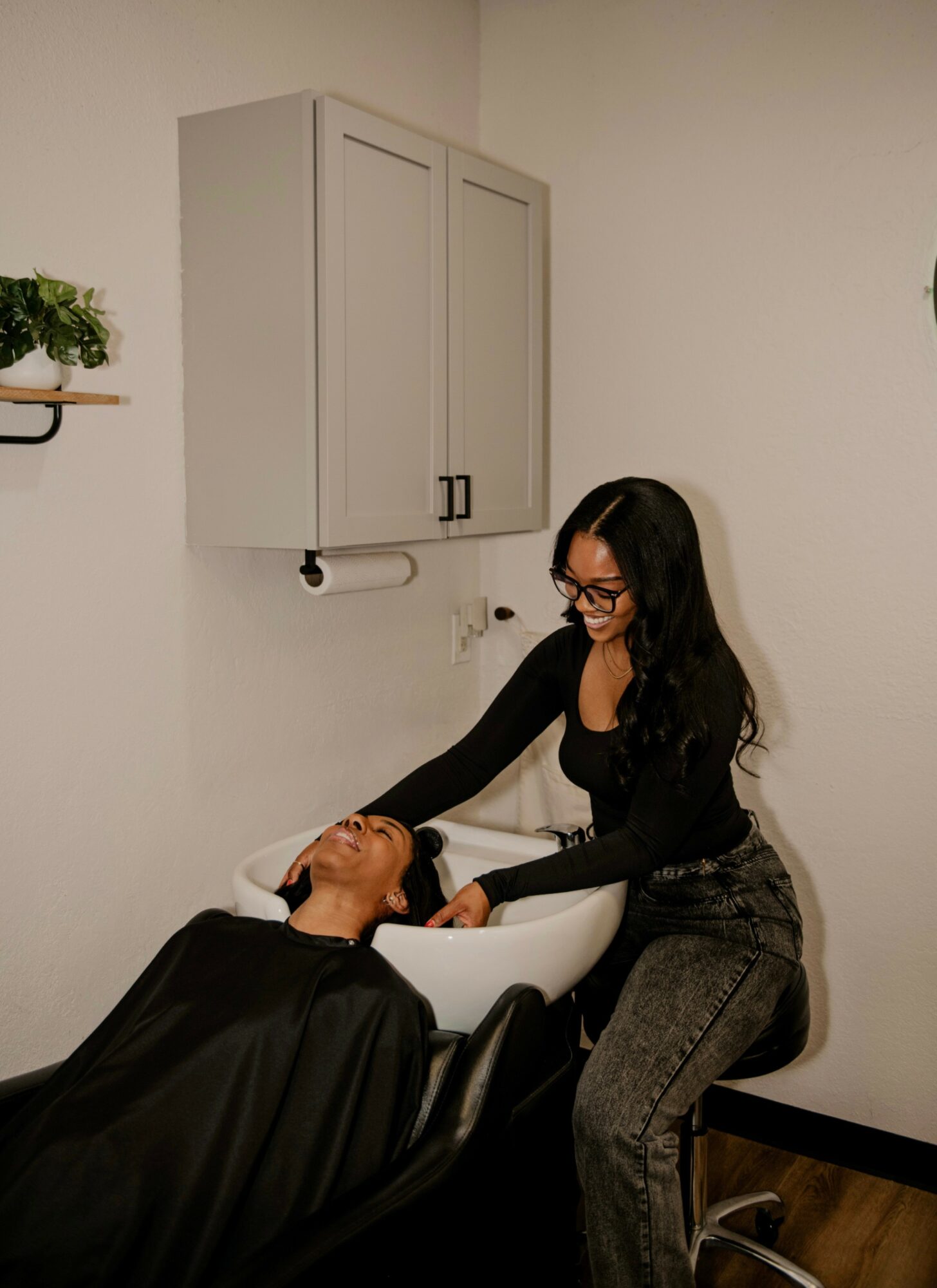 Woman washing a person's hair in a salon sink, smiling, with a white wall and cabinet in the background.