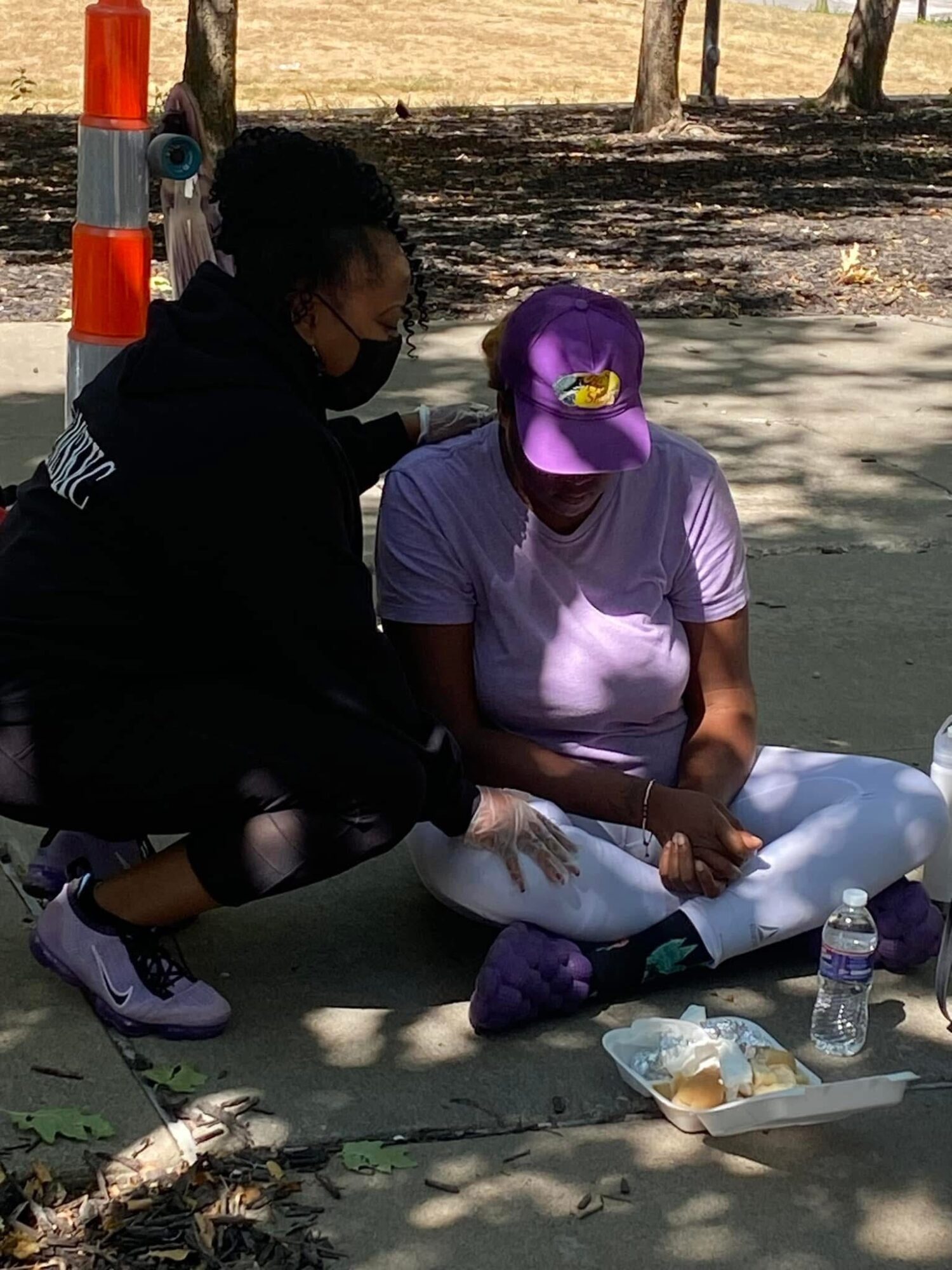 Two women sitting on the ground outdoors, one wearing a purple cap and white shirt, the other in black, with food and water bottles nearby.