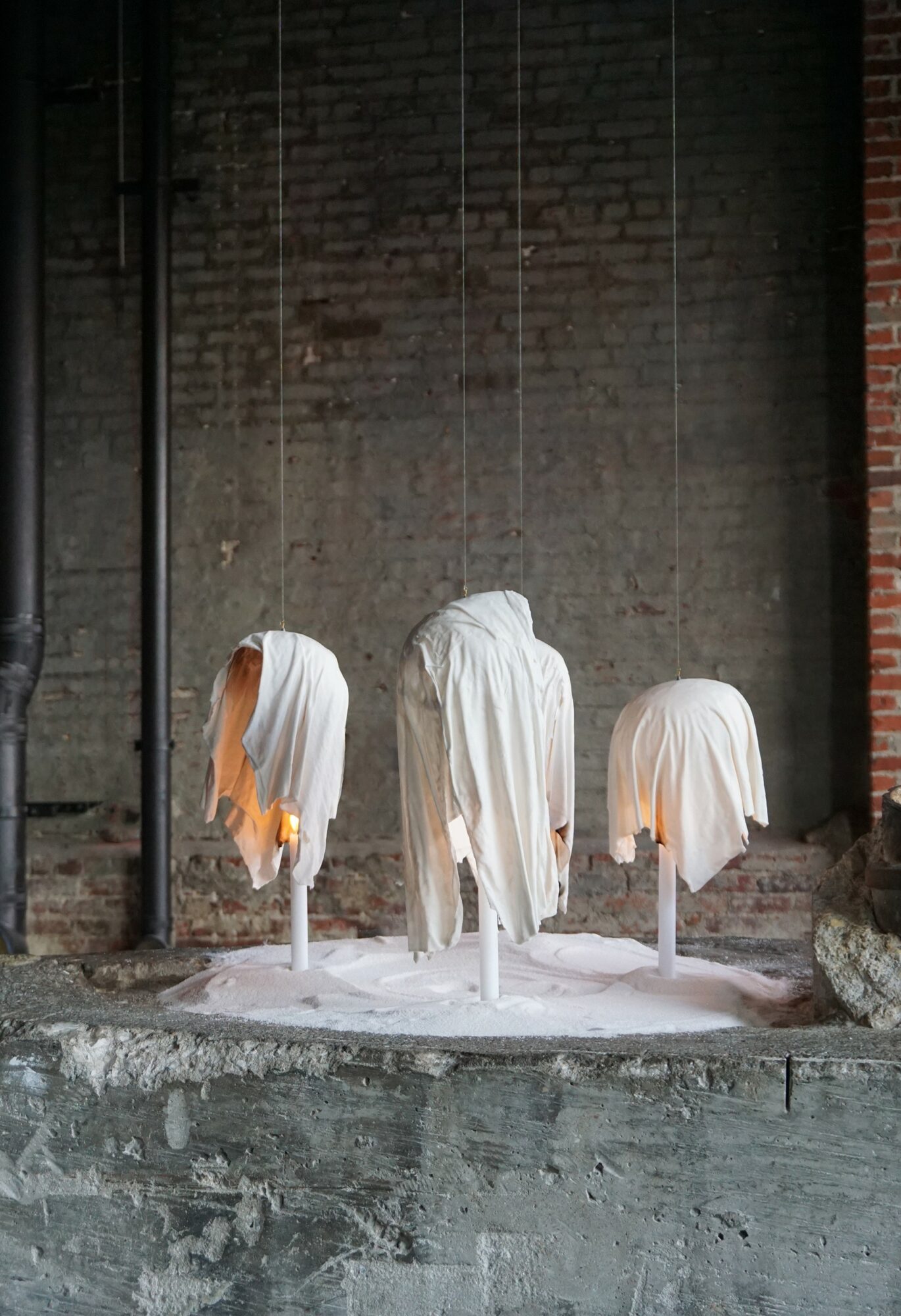 Three ghostly figures draped in white cloths stand on a circular platform against a brick wall.