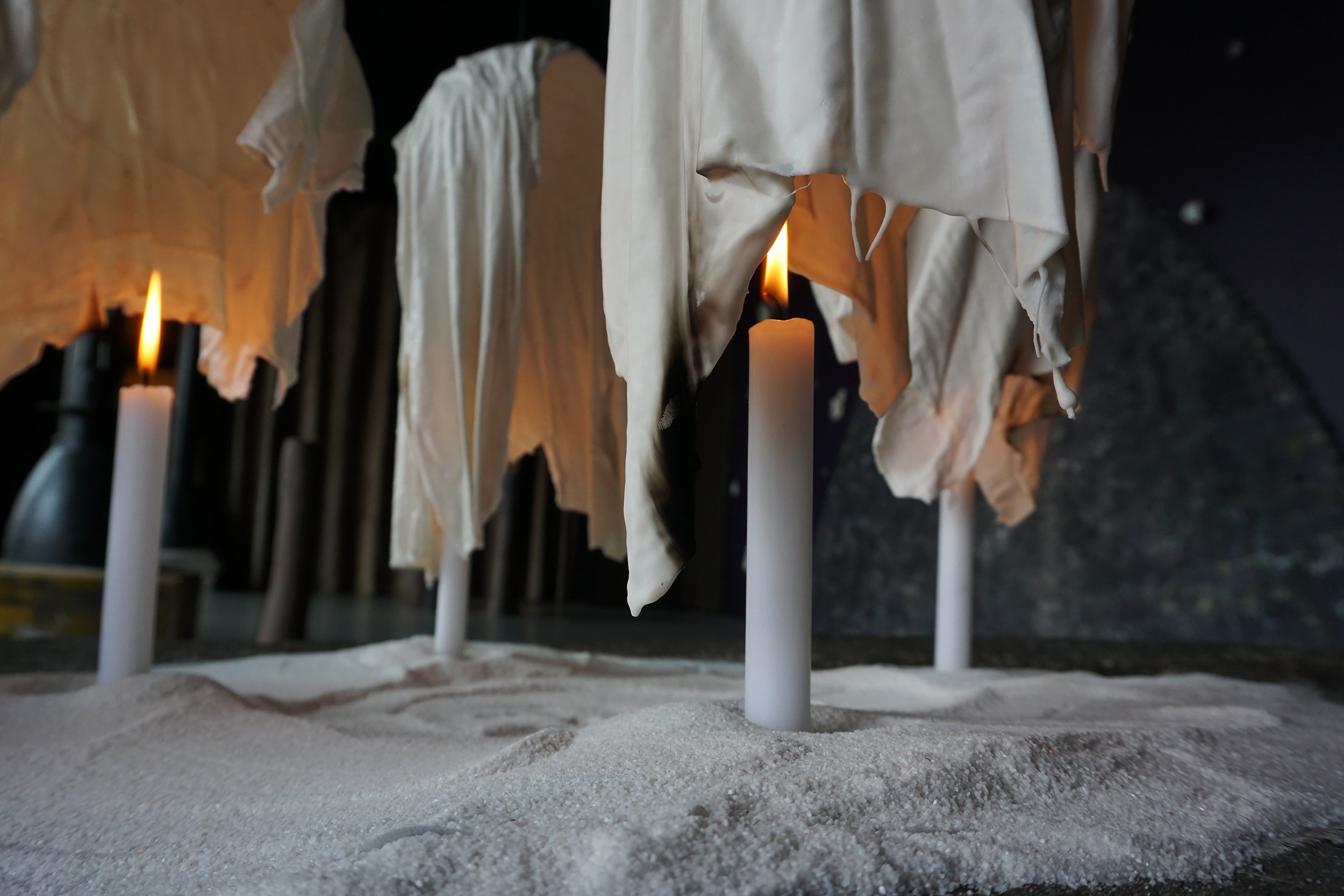 Clothes hanging on poles above sand, with candles burning nearby at night.