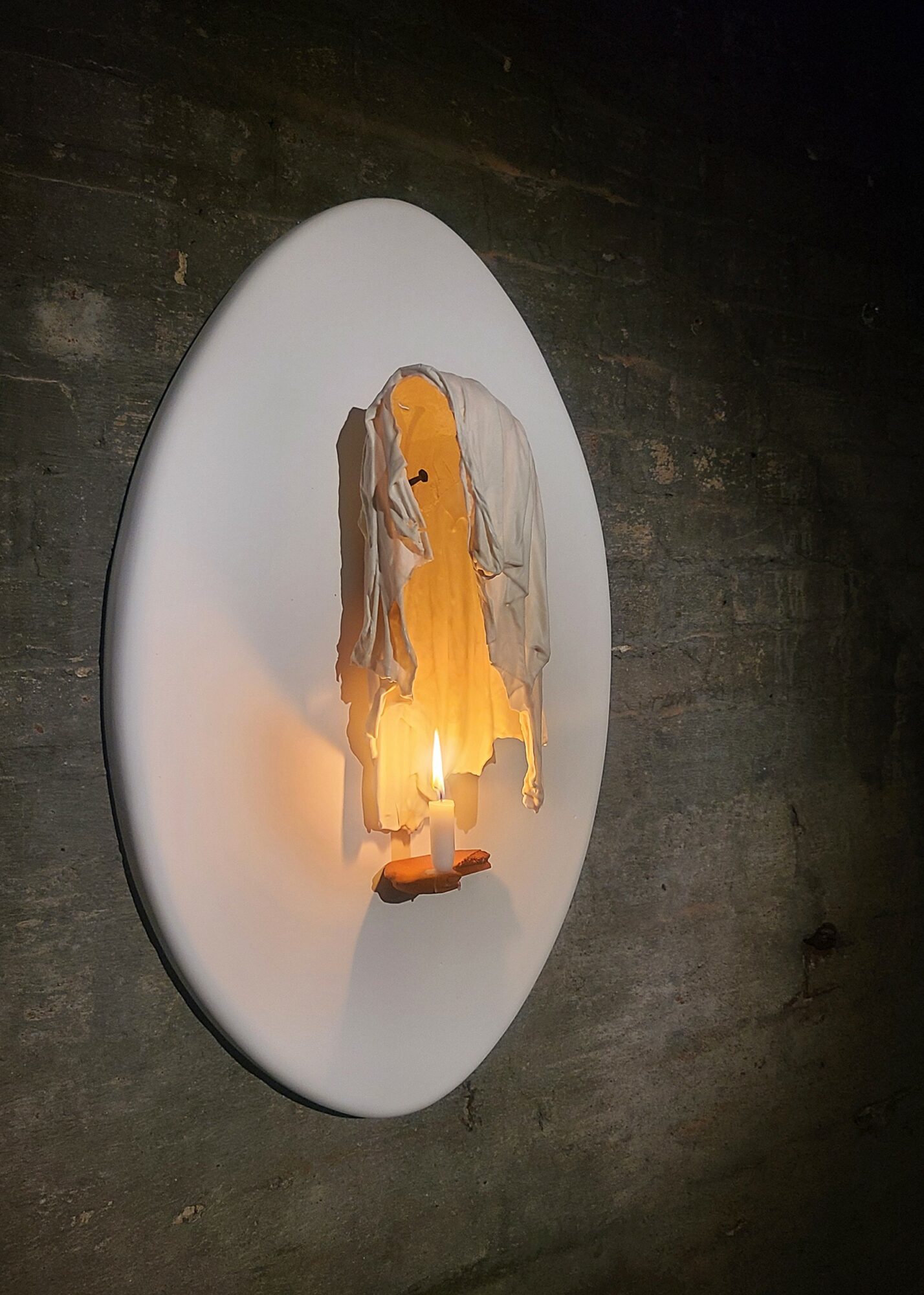 Sculpture of a figure with a yellow face, gray hair, and a white dress, illuminated from below, mounted on a white oval background.