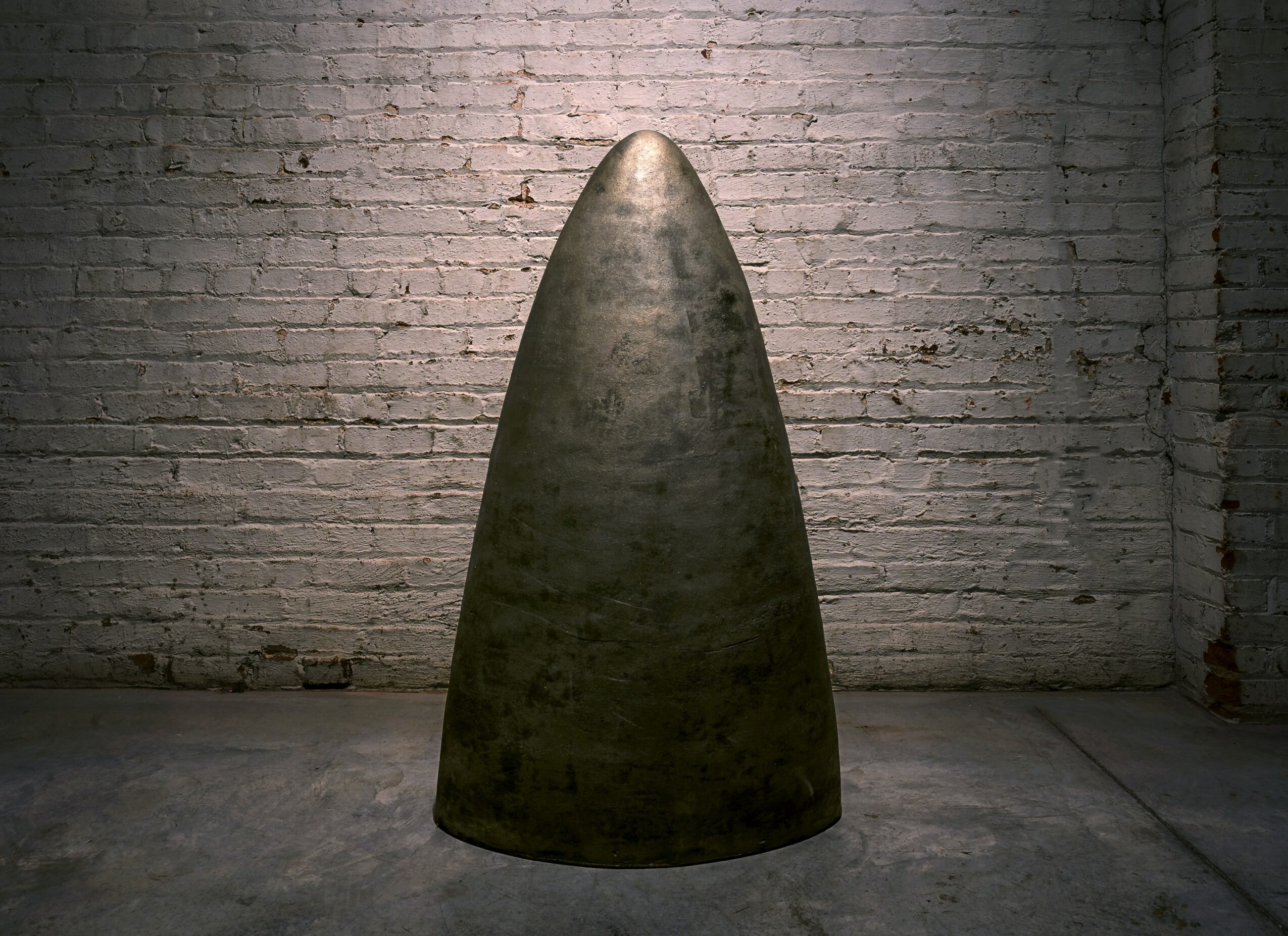A large, cone-shaped object stands in front of a brick wall in a dimly lit space.