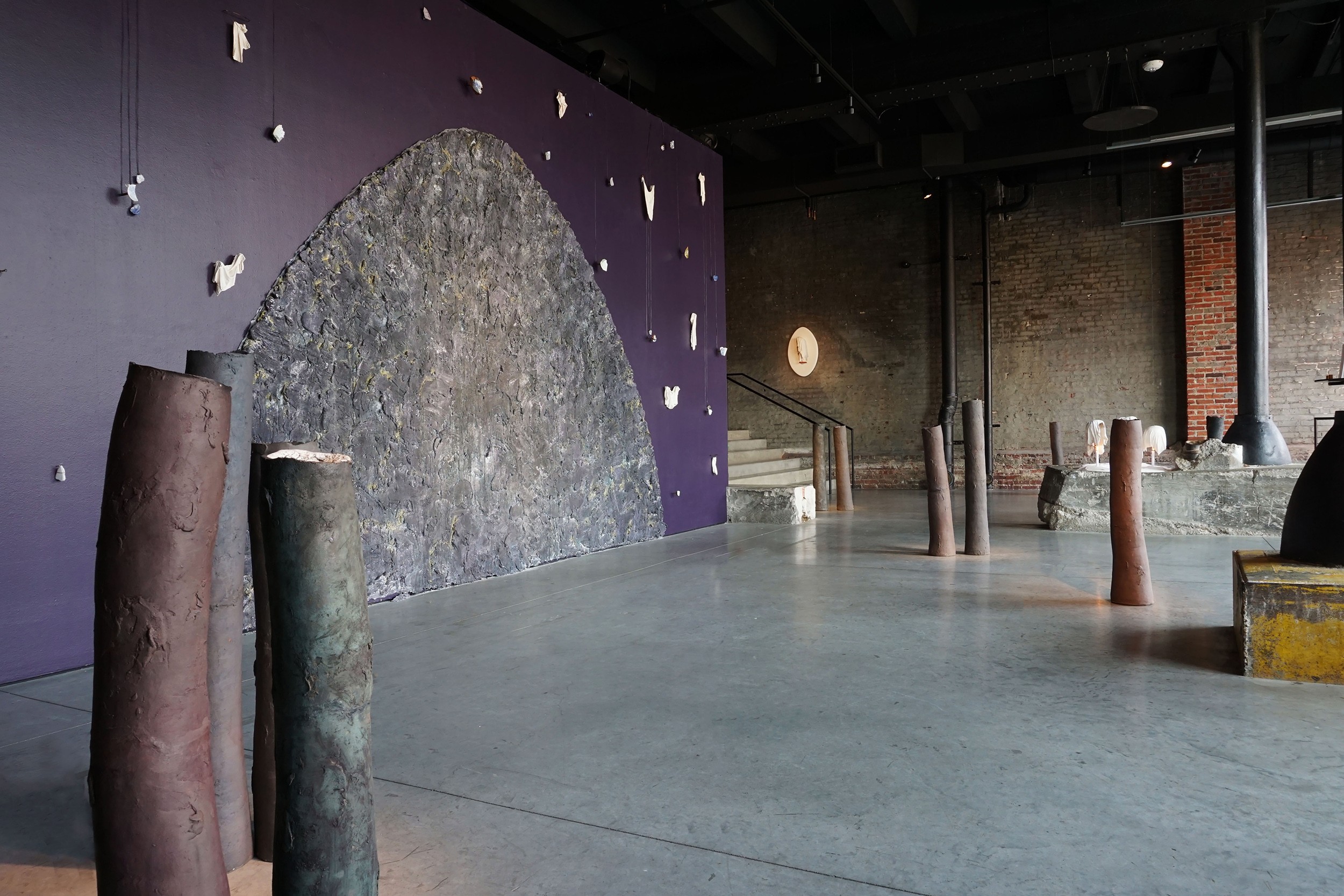 Indoor space with a purple wall, large textured gray arch, and various cylindrical objects on the floor.