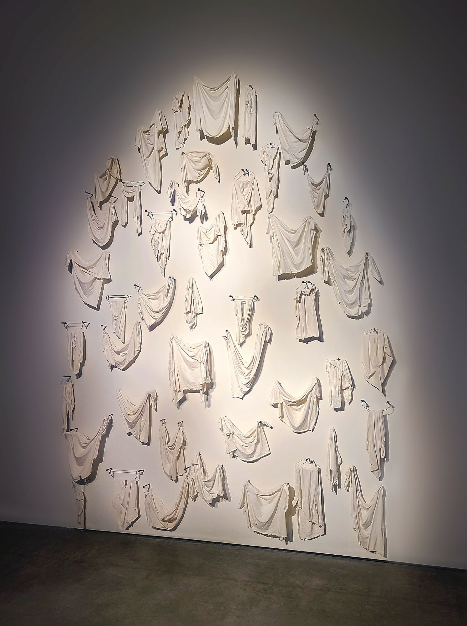Wall installation of multiple white fabric pieces hanging in a pattern, illuminated by a spotlight, on a gray wall.