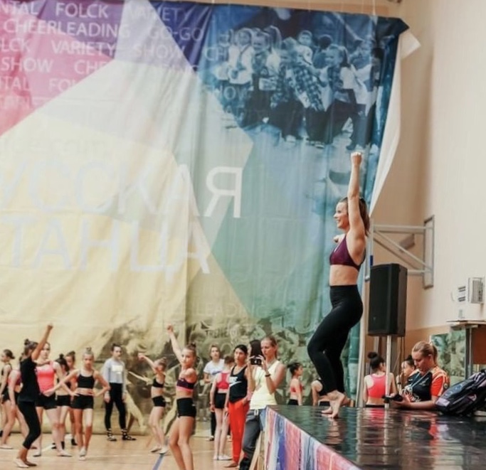 A group of dancers performing on stage with a colorful backdrop and audience watching, some dancers raising arms.