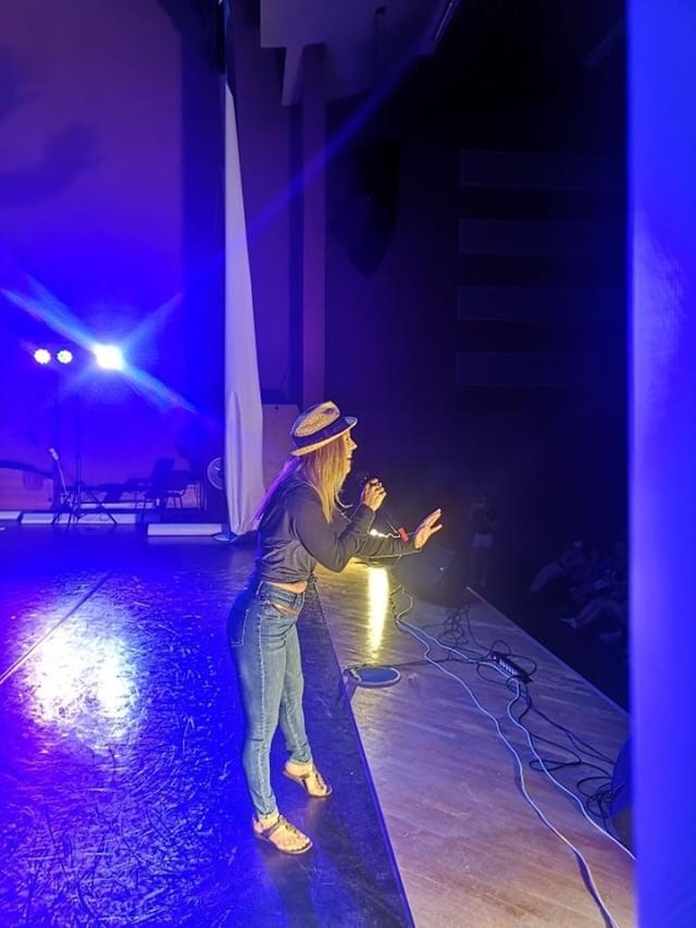 Person wearing a hat and jeans speaking into a microphone on stage with blue lighting.