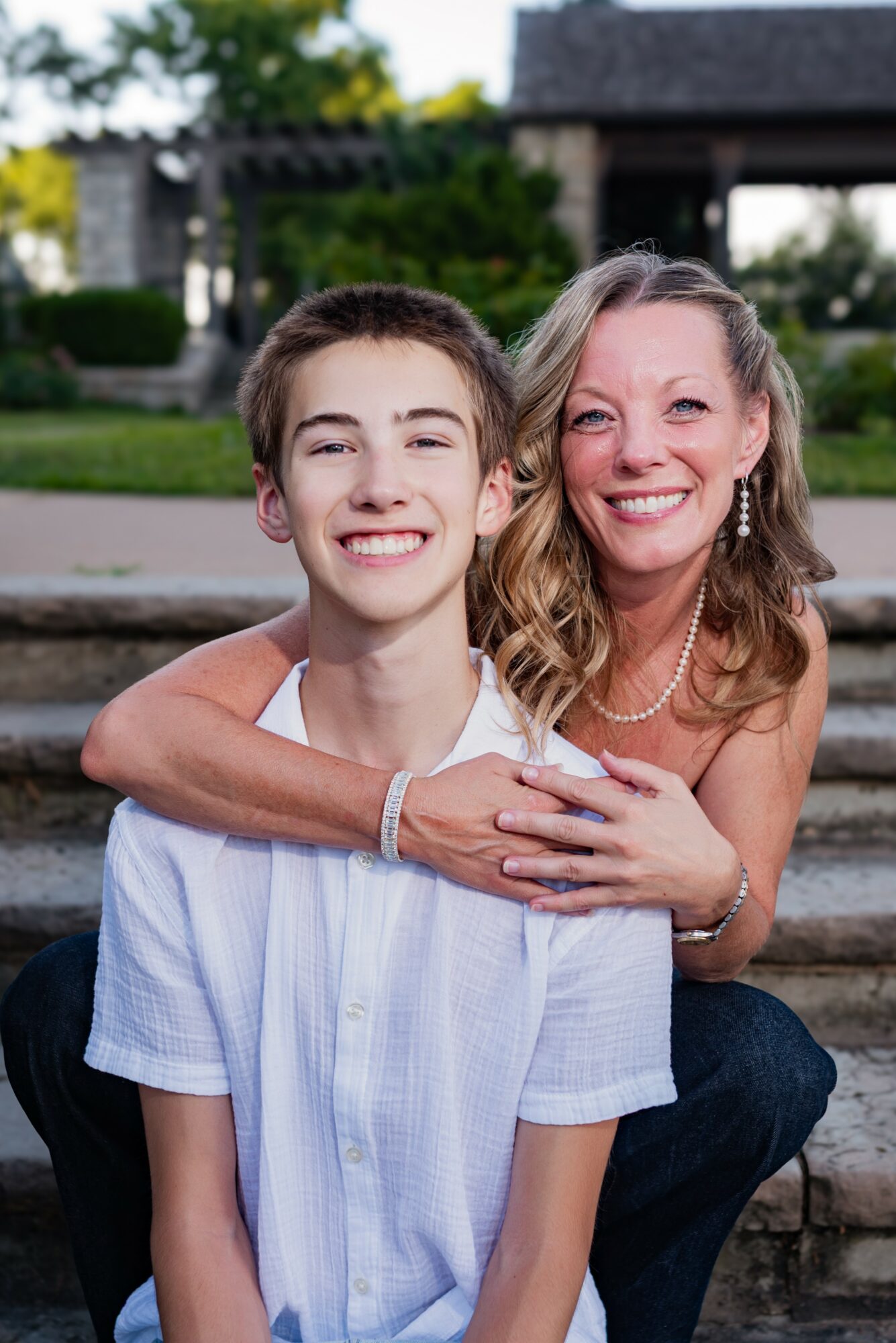 A woman and a young boy smile and hug outdoors, with a house and greenery in the background.