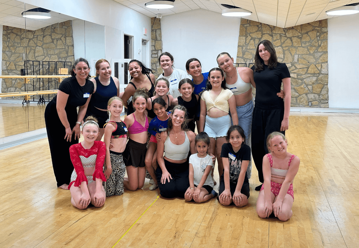 Group of young girls and women in dance studio, some in dance attire, posing together indoors.