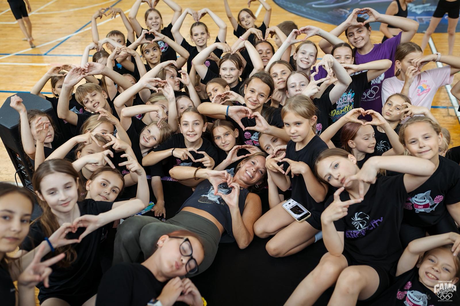 Group of young girls and women in a gymnasium, making heart shapes with their hands, smiling and posing together.