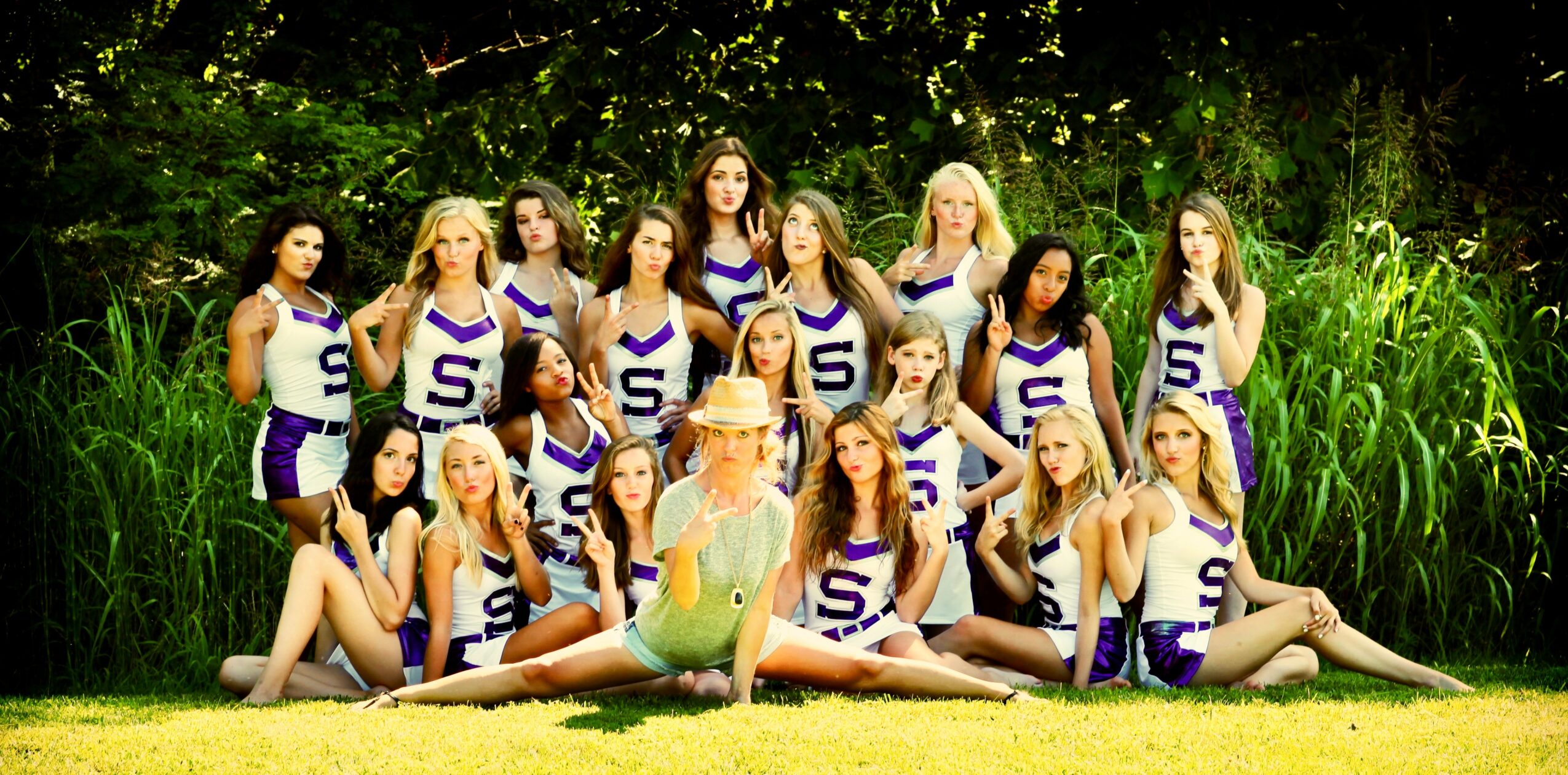 Group of young women in cheerleading uniforms with a person in a bird costume outdoors, surrounded by greenery.