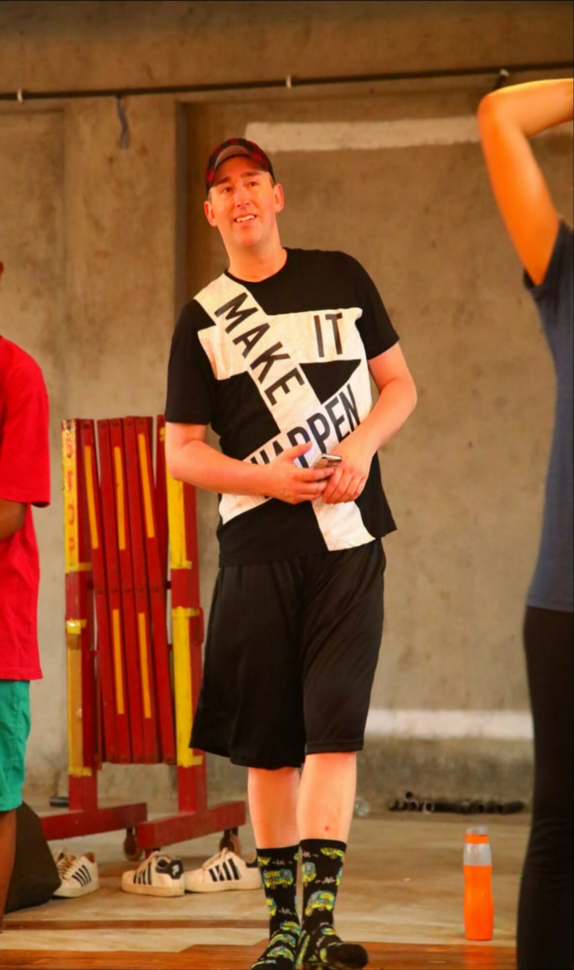 Person standing indoors, smiling, wearing a black and white shirt, black shorts, and colorful socks, with a concrete wall background.