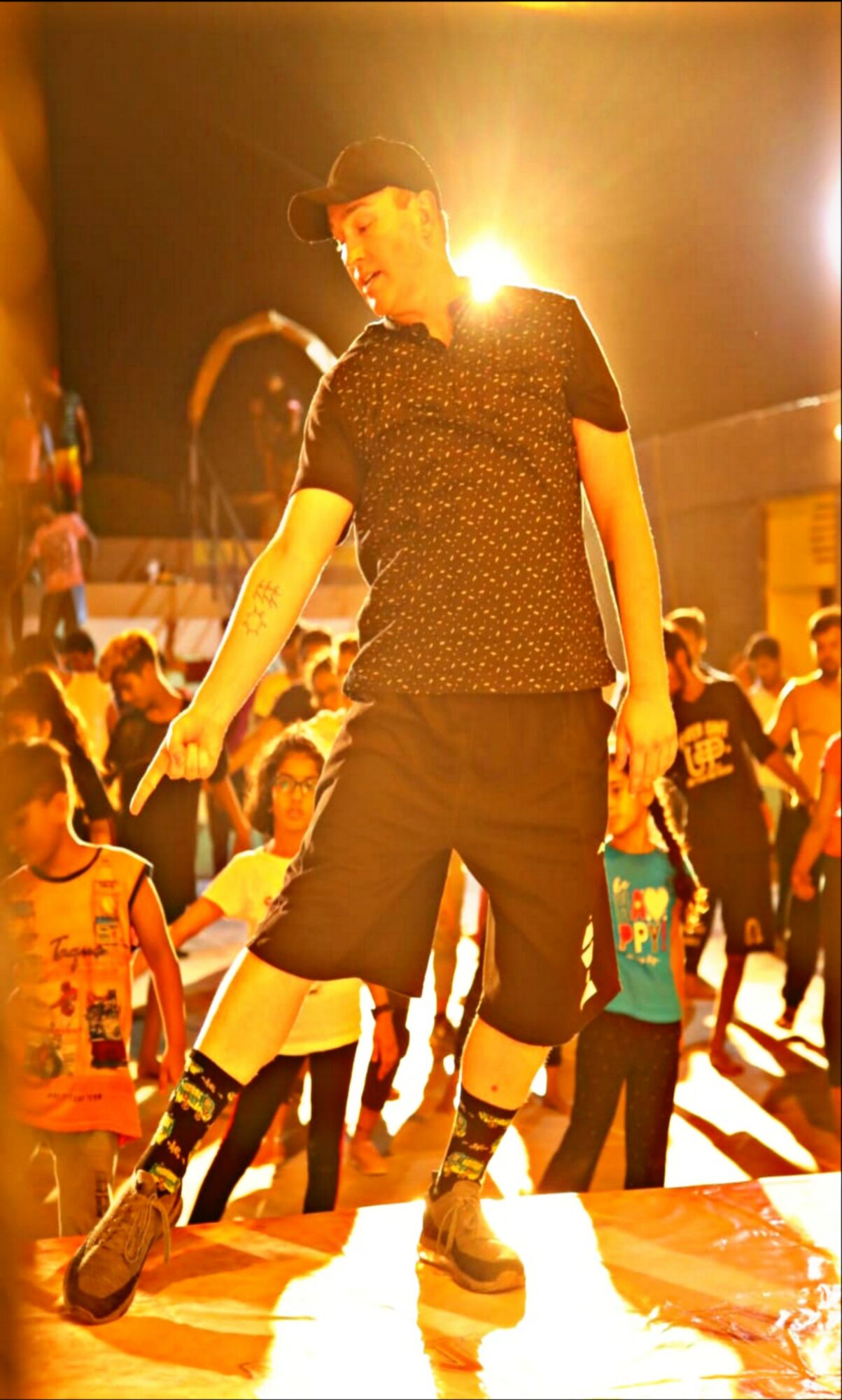 Person dancing on stage with a crowd in the background, bright stage lighting, wearing a hat, black shirt, and shorts.
