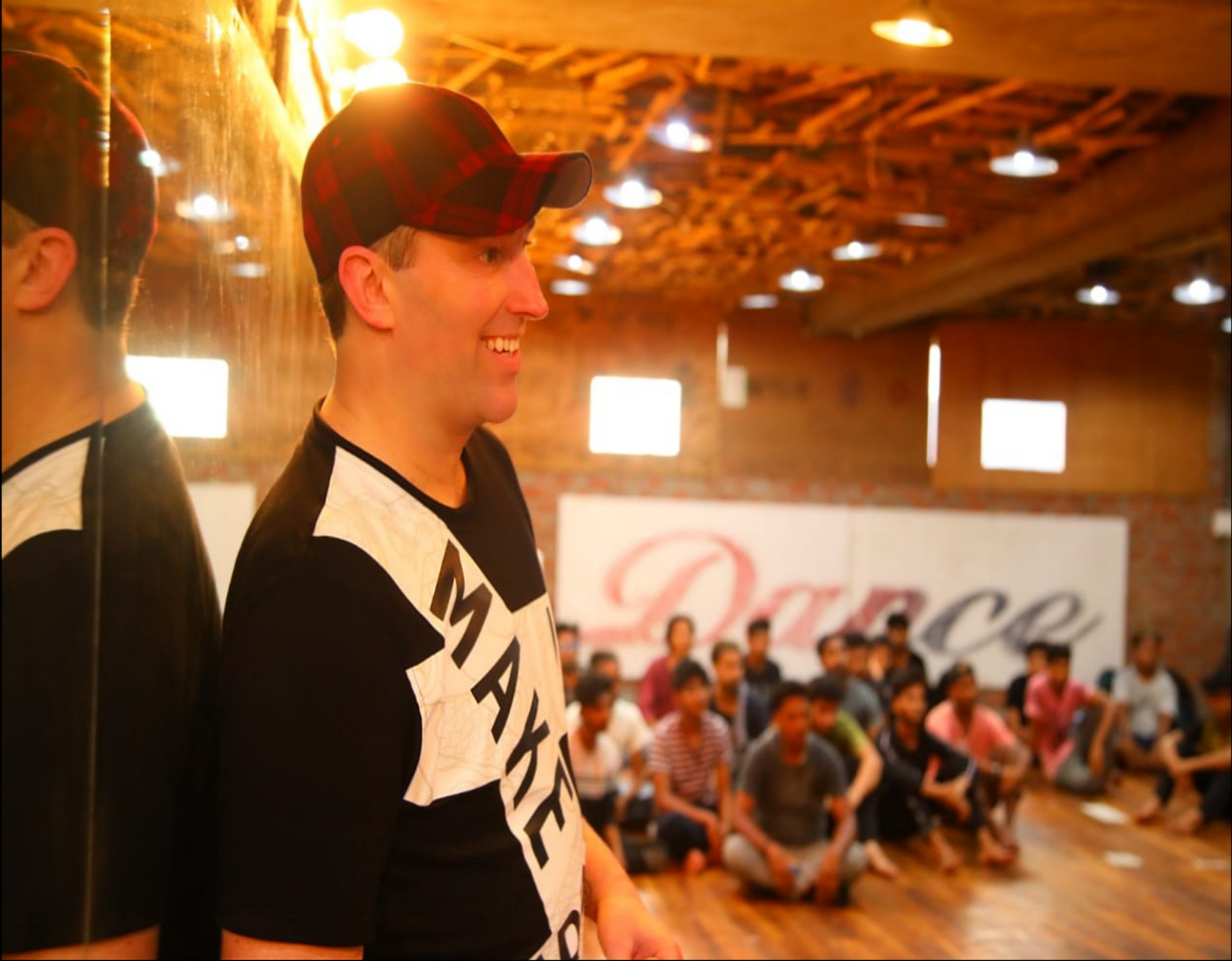 Young man in a black and white shirt and cap smiling, with a group of people sitting on the floor in the background.