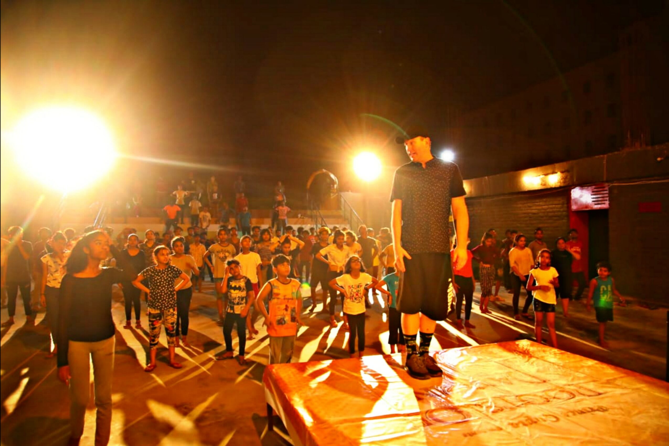 Children and adults gather in a gymnasium with bright lights, some standing on a stage, others watching or participating.