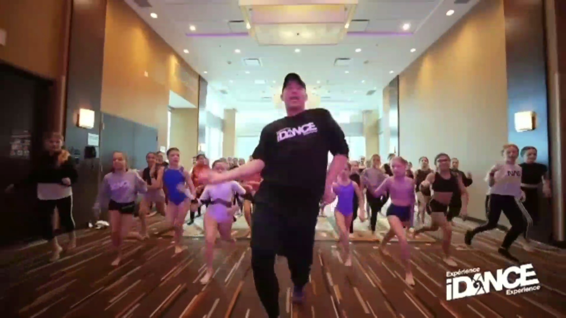 Group of people dancing in a spacious room with a person leading in the center, wearing a black shirt with 'DANCE' written on it.