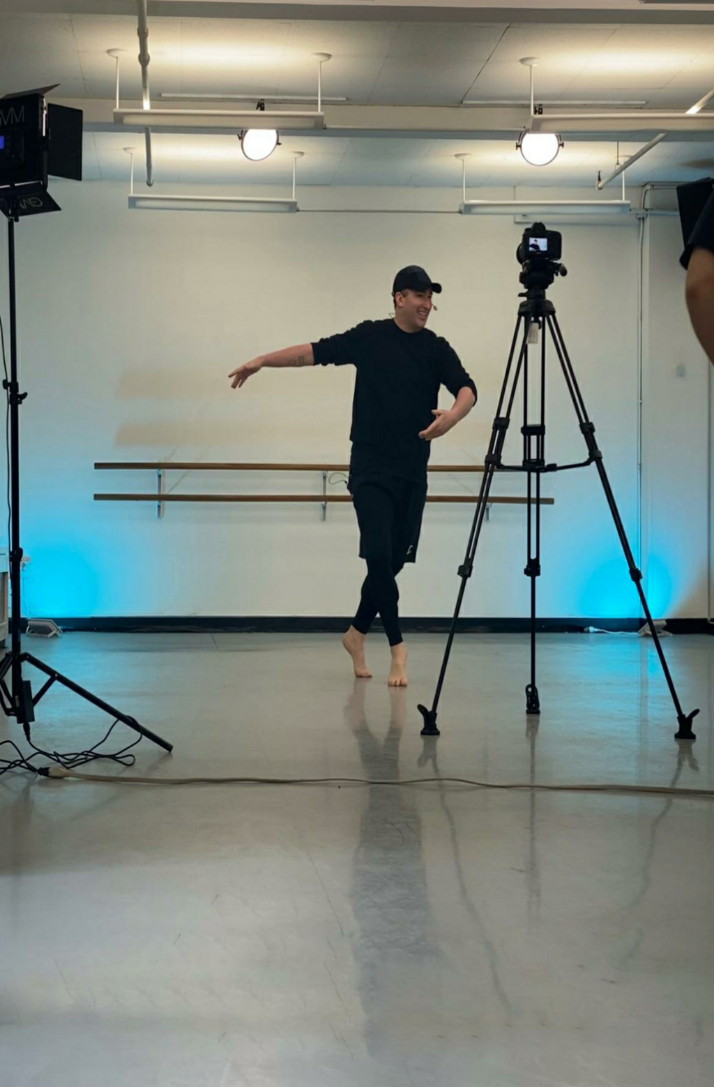 Person in black clothing and cap standing in a dance studio with camera equipment, blue lighting on wall, wooden ballet barres