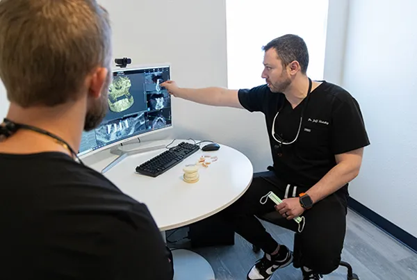 Two medical professionals in black scrubs discuss brain scans in a clinical setting, one pointing at the monitor. Word count: 20