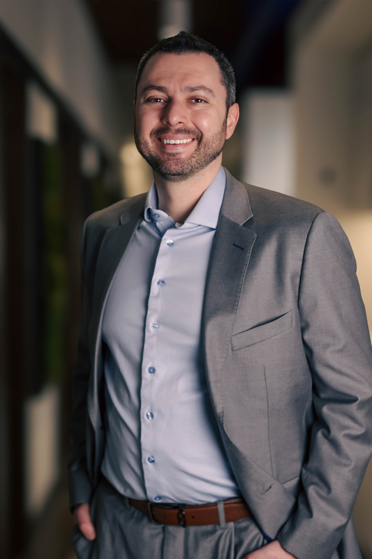 Man in gray suit and light blue shirt smiling indoors with blurred background.
