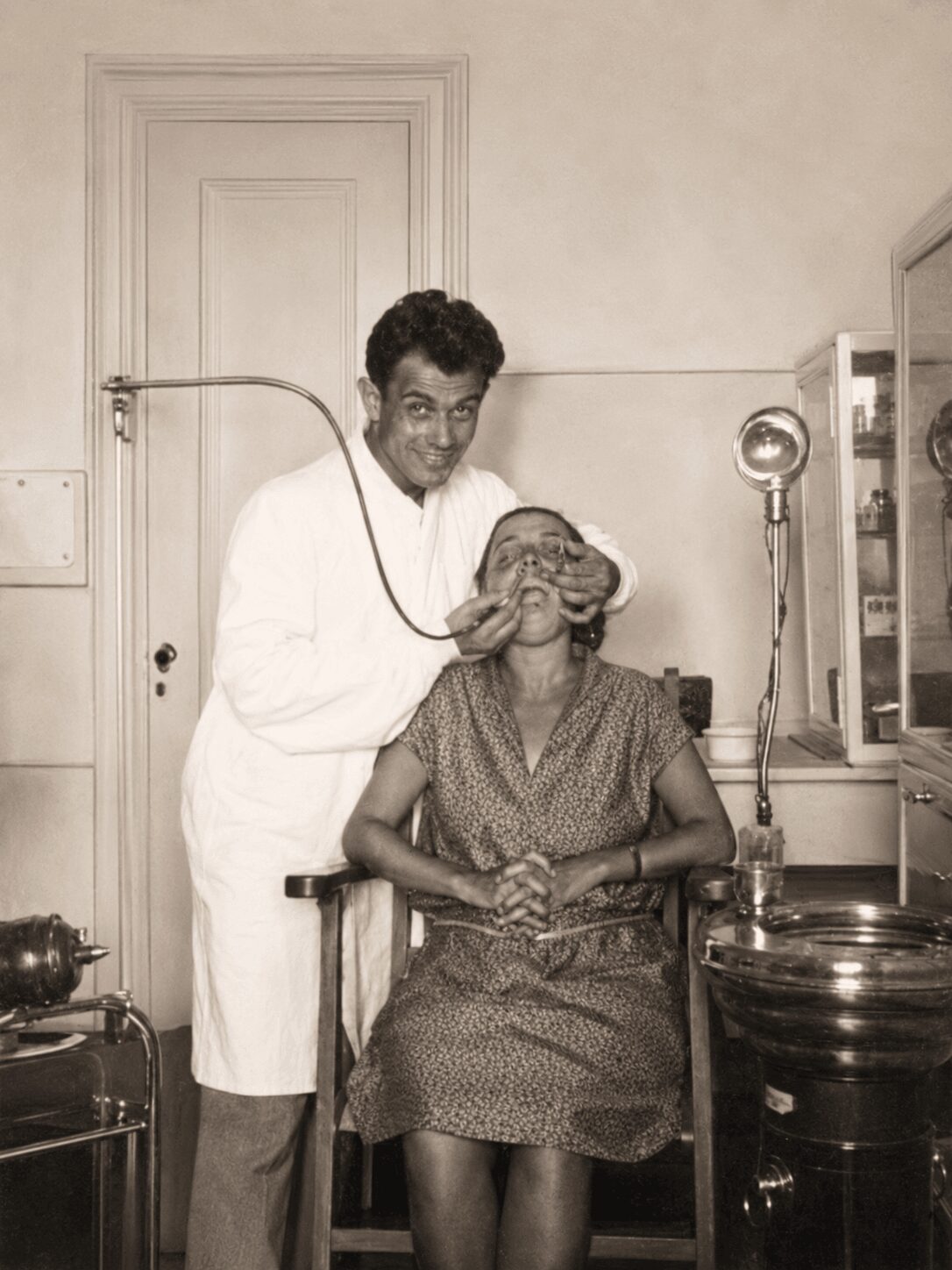 A man in a white coat stands behind a seated woman in a dress, holding her head gently in a medical setting.