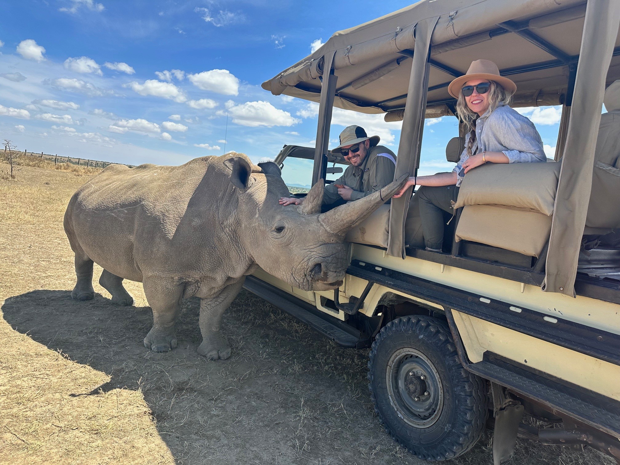 Two people in a safari vehicle interacting with a rhinoceros in a dry, open landscape under a partly cloudy sky.