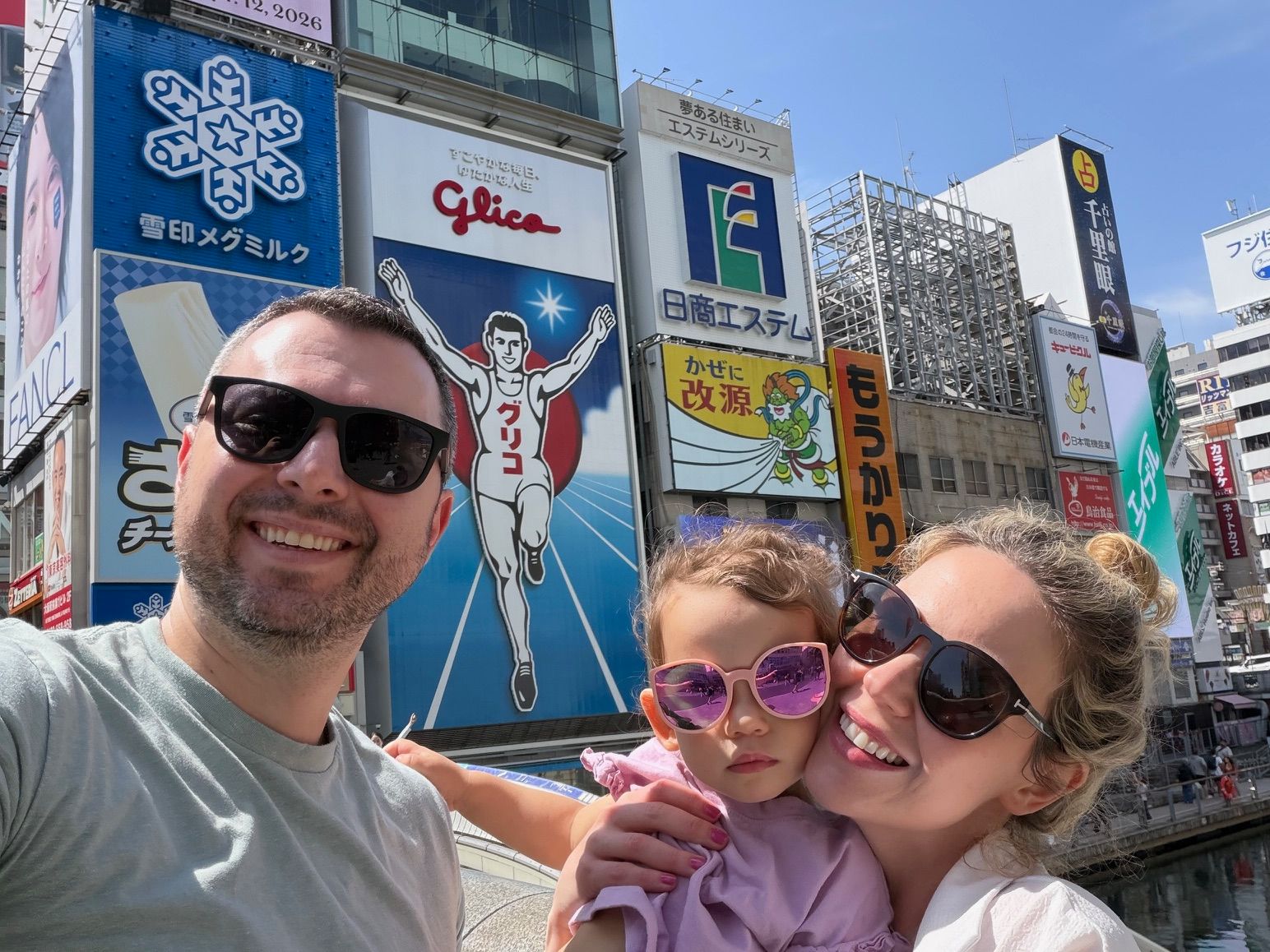 Three people smiling in front of colorful billboards and signs in an urban area, with a clear blue sky above.