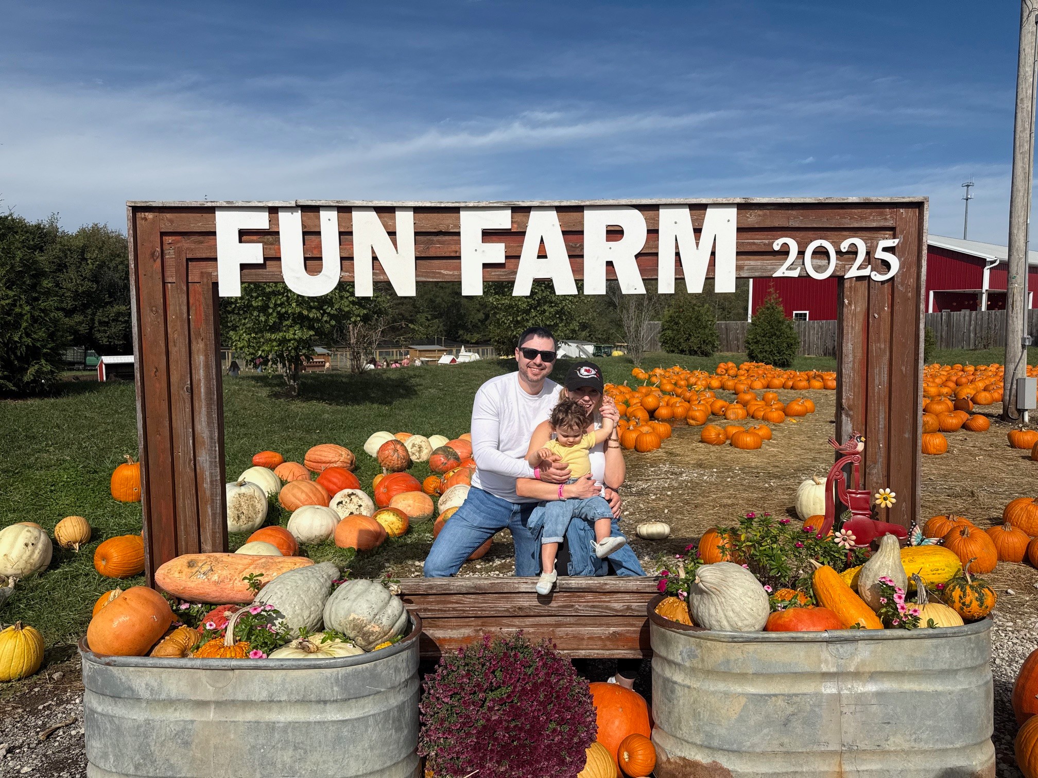 Two people sitting on a bench at a pumpkin patch with pumpkins around them and a sign reading 'FUN FARM 2025'.