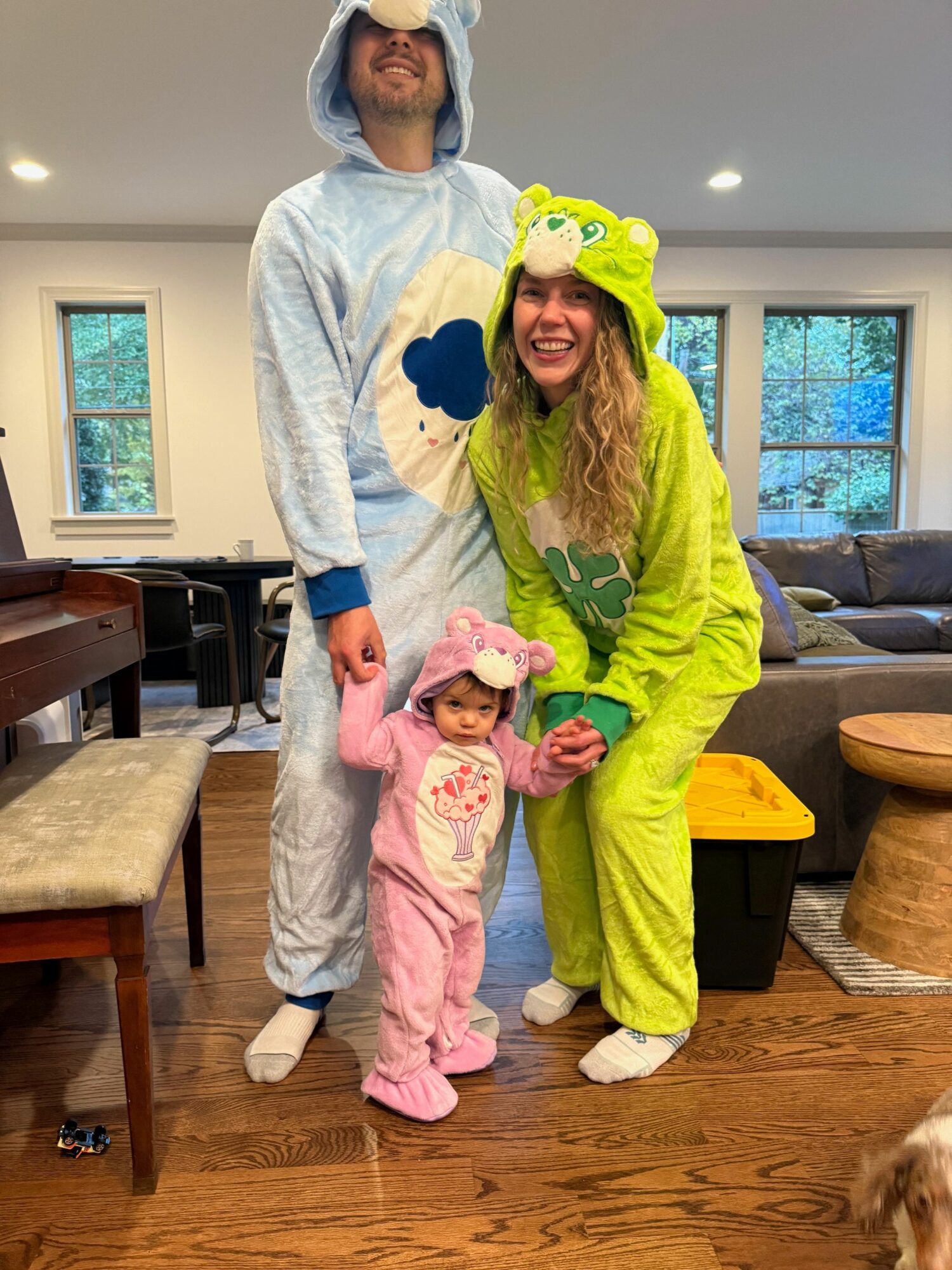 Two people in colorful costumes with a child in pink pajamas, indoors near a window and wooden furniture.